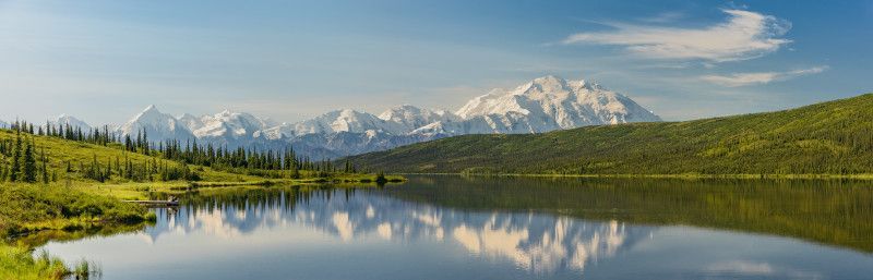 Snowy mountains reflected in a calm lake under a blue sky, with green forested shores.