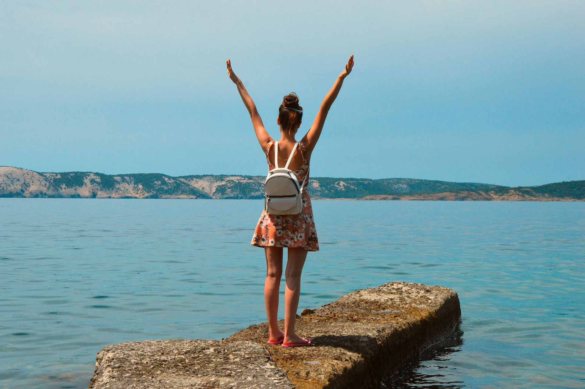 Person standing on a rocky pier with arms raised, facing a calm blue sea and distant coastline