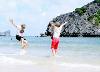 Two people jump in shallow beach water near a rocky cliff under a bright sky