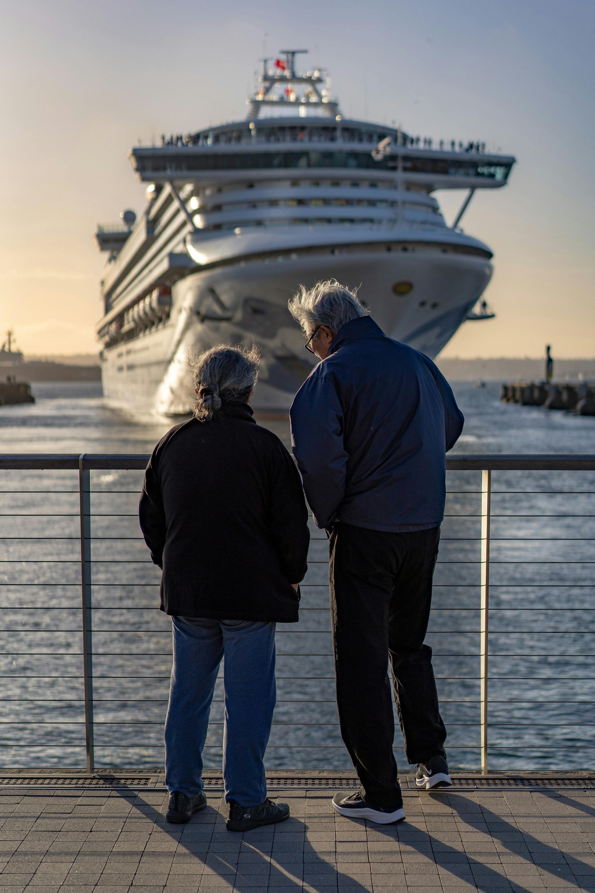 Two people stand by a railing watching a large cruise ship approach the harbor.