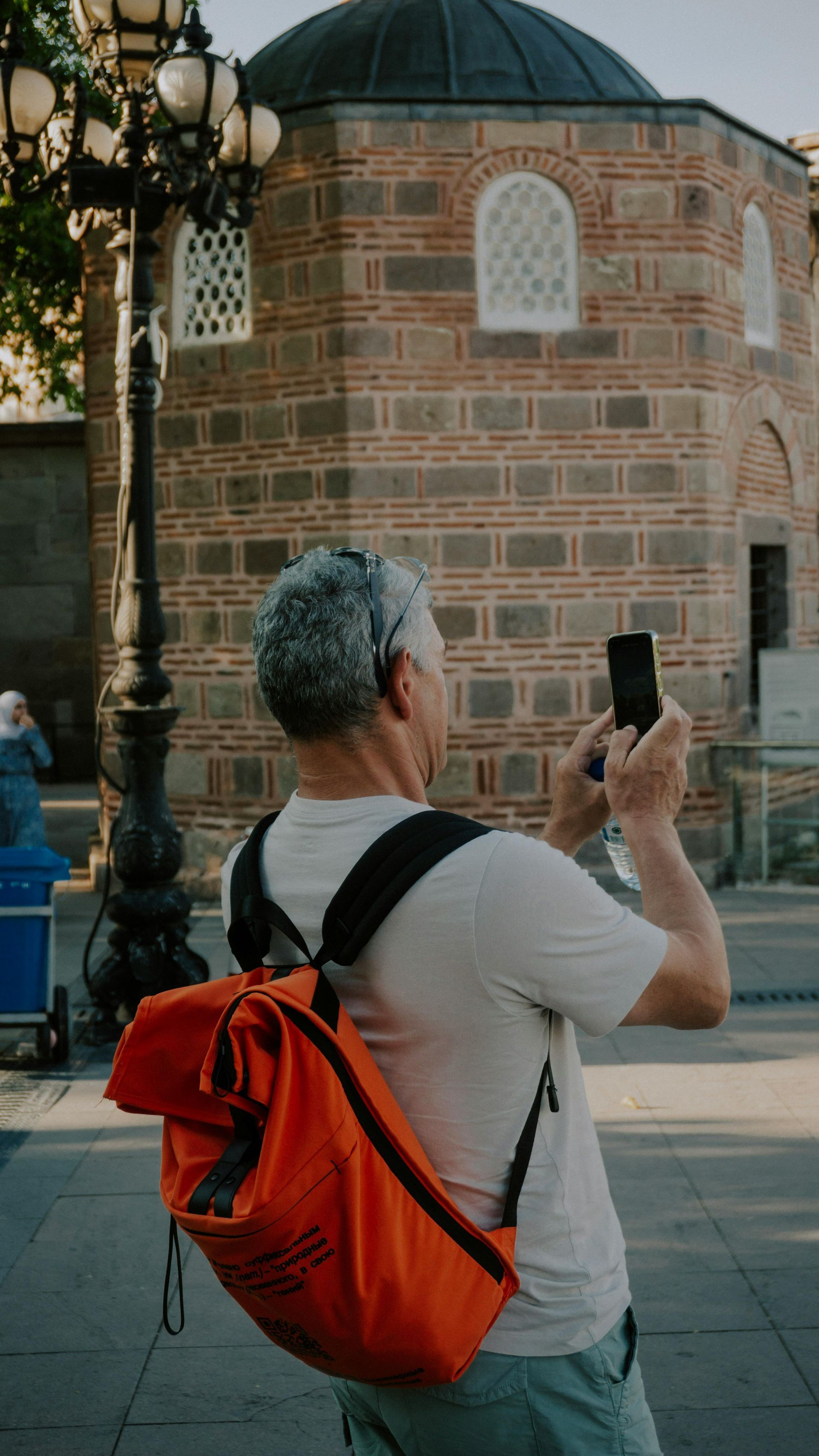Person with orange backpack photographing a historic brick building and lamp post in a city square