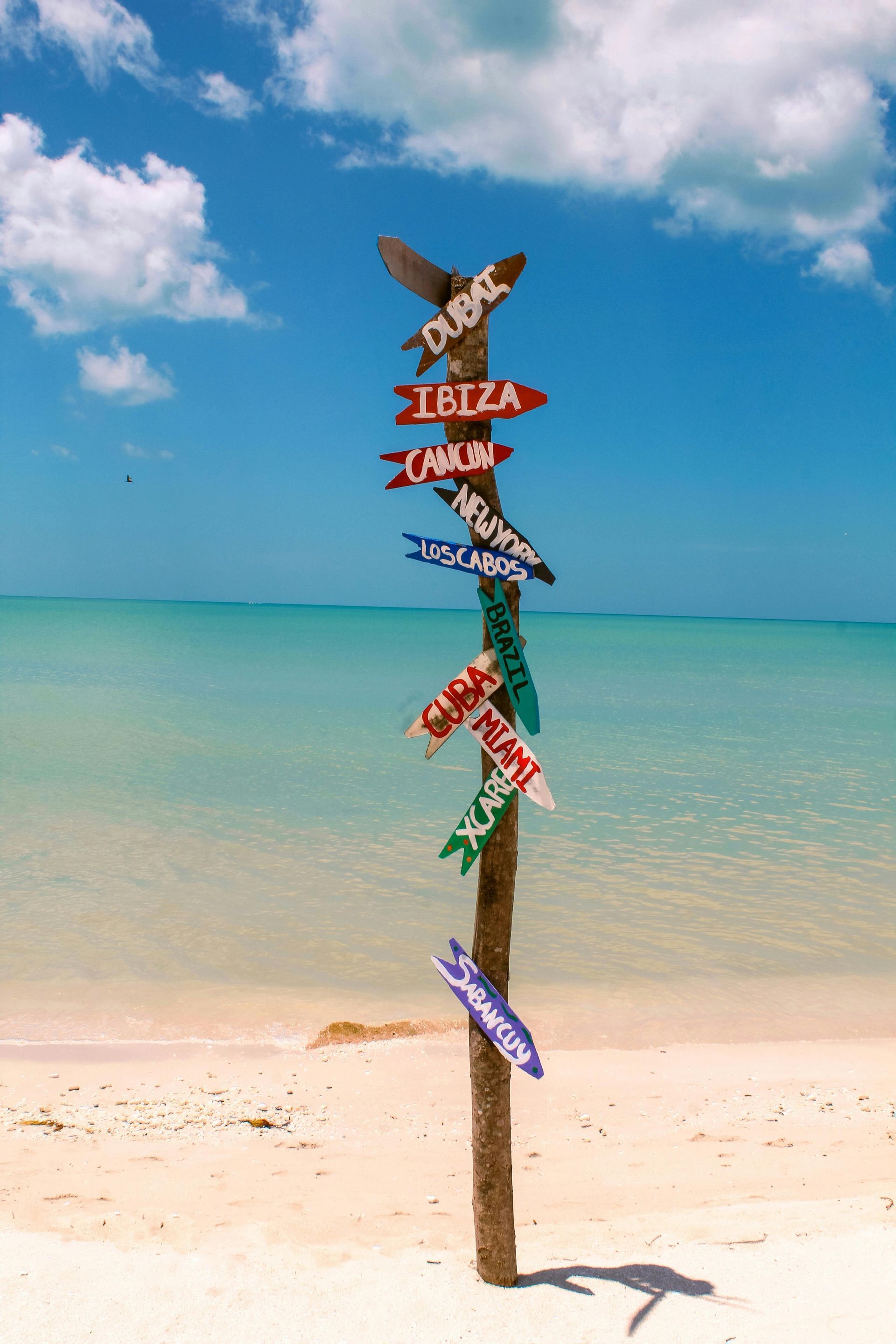 Beach signpost with colorful directional arrows on a sandy shore under a bright blue sky