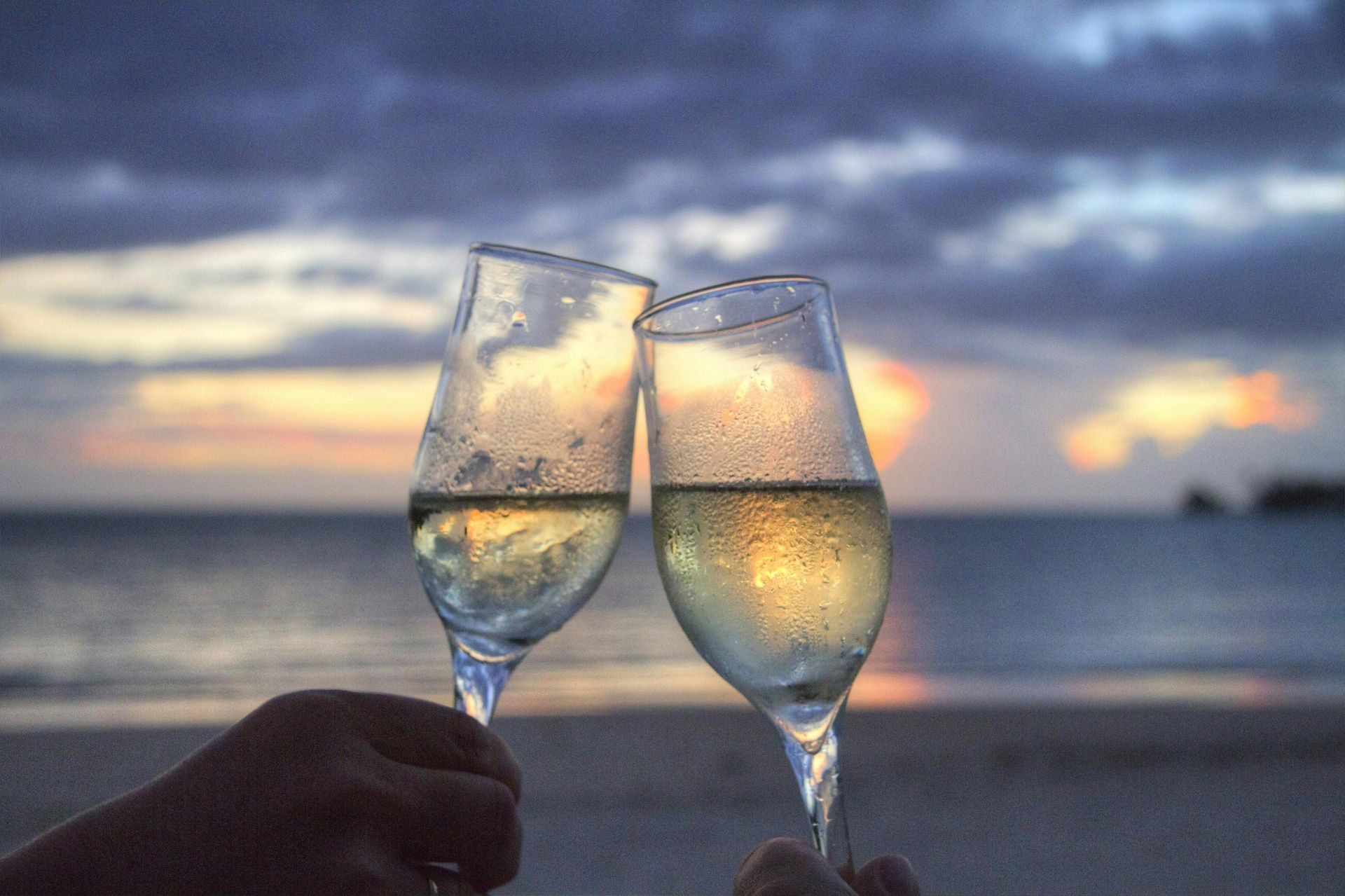 Two champagne flutes toasting at sunset on a beach, with ocean and cloudy sky in the background