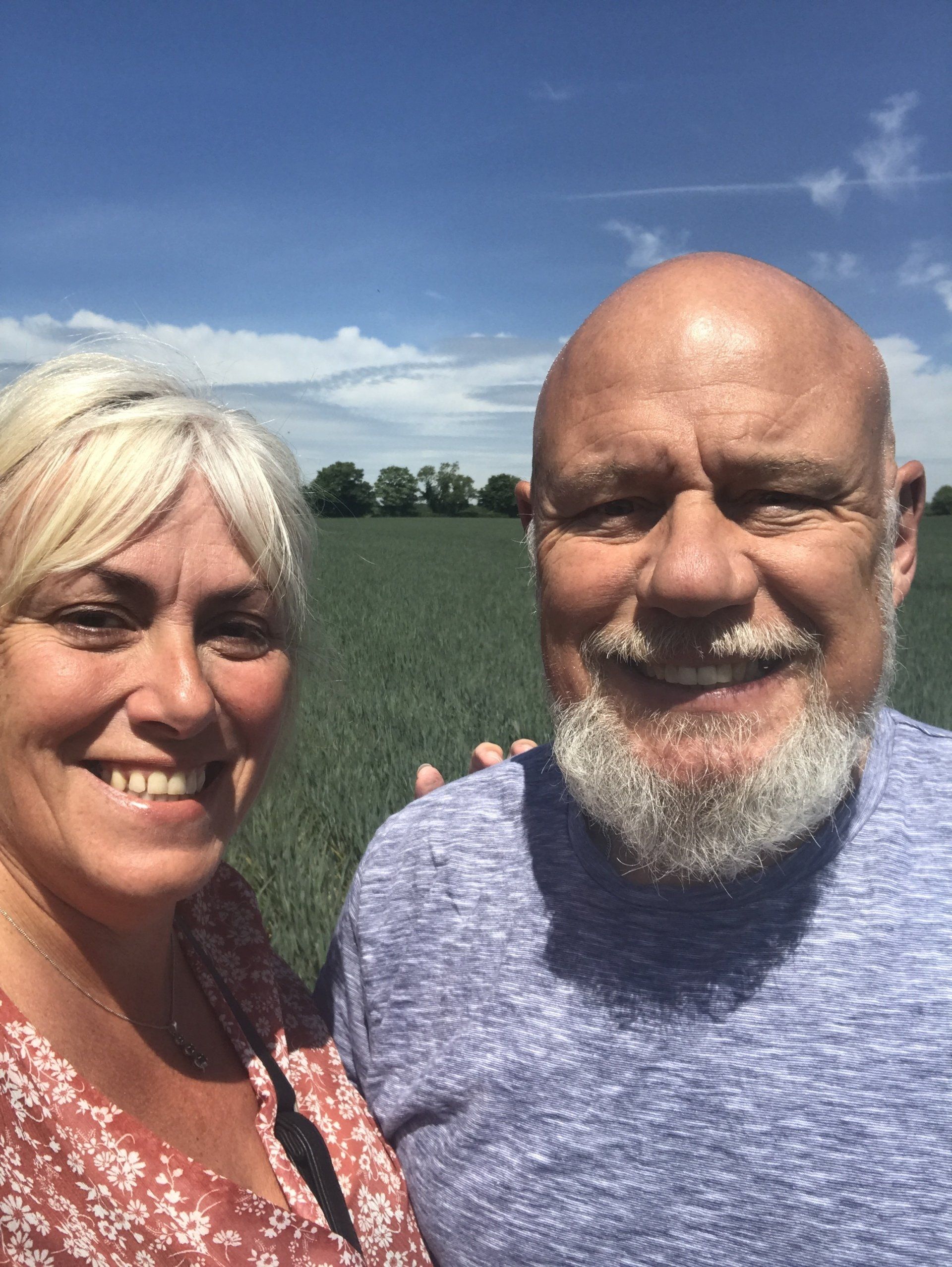 Woman and man smiling for a selfie, standing in a field on a sunny day.