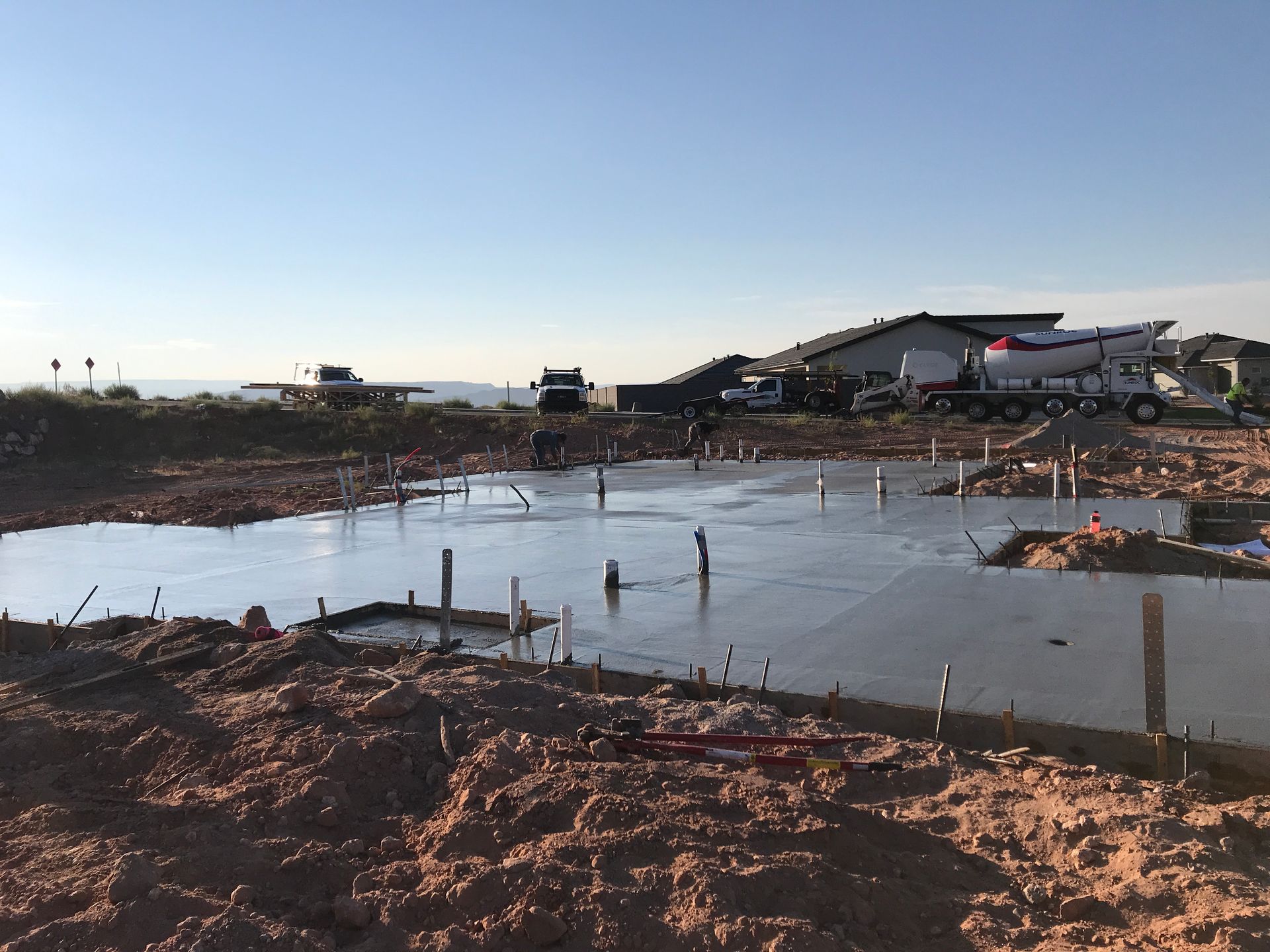 Shallow muddy water at a construction site with rubble, stakes, and trucks under a clear sky.