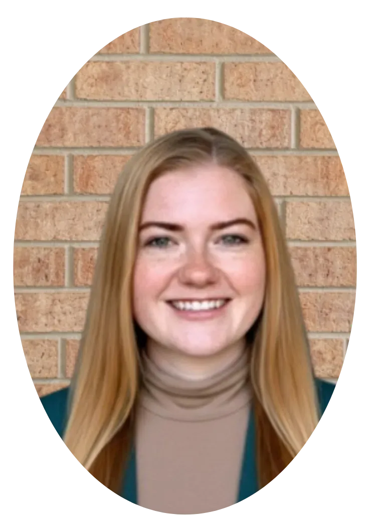 A person with long blonde hair and a friendly smile, wearing a beige turtleneck and green jacket against a brick wall.