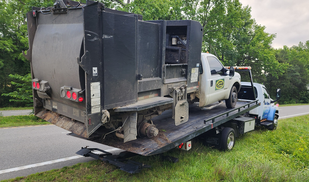 A white garbage truck is loaded on a tow truck, roadside. Dark grey body is damaged | Chip Ragans Auto, LLC