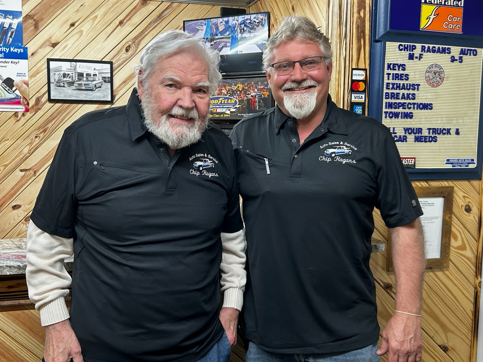 Two men with beards pose in a garage. Both wear black shirts, smiling. Behind them, a sign lists auto services | Chip Ragans Auto, LLC