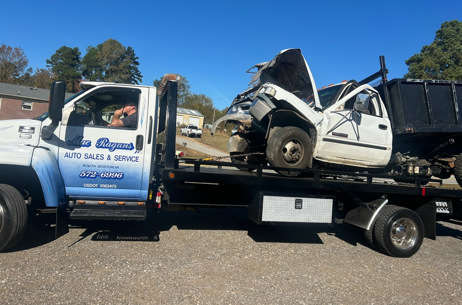 Tow truck carrying a severely damaged white truck. The tow truck has blue graphics. Outdoors on a sunny day | Chip Ragans Auto, LLC