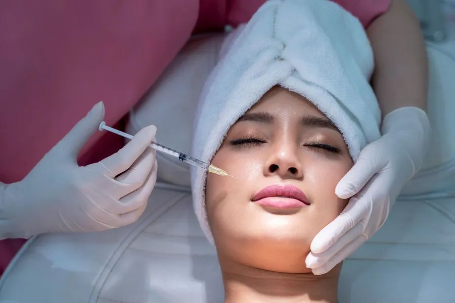 Woman receiving facial injection with needle; medical setting, hands in gloves.