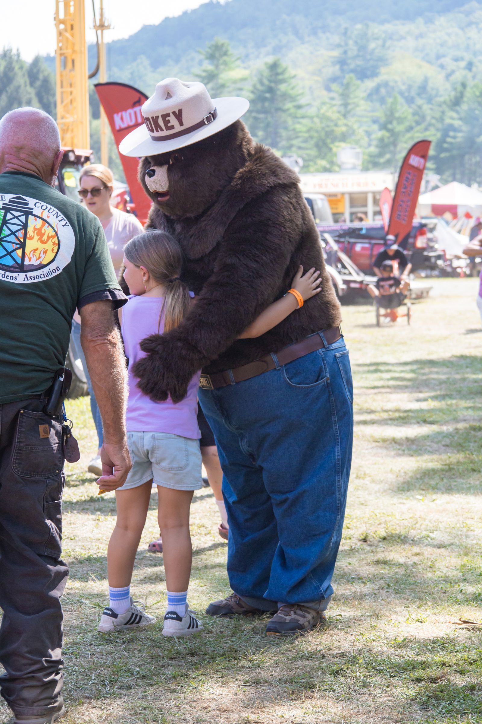 Person in a cowboy hat hugging a child outdoors at a fair with red flags and mountains in the background
