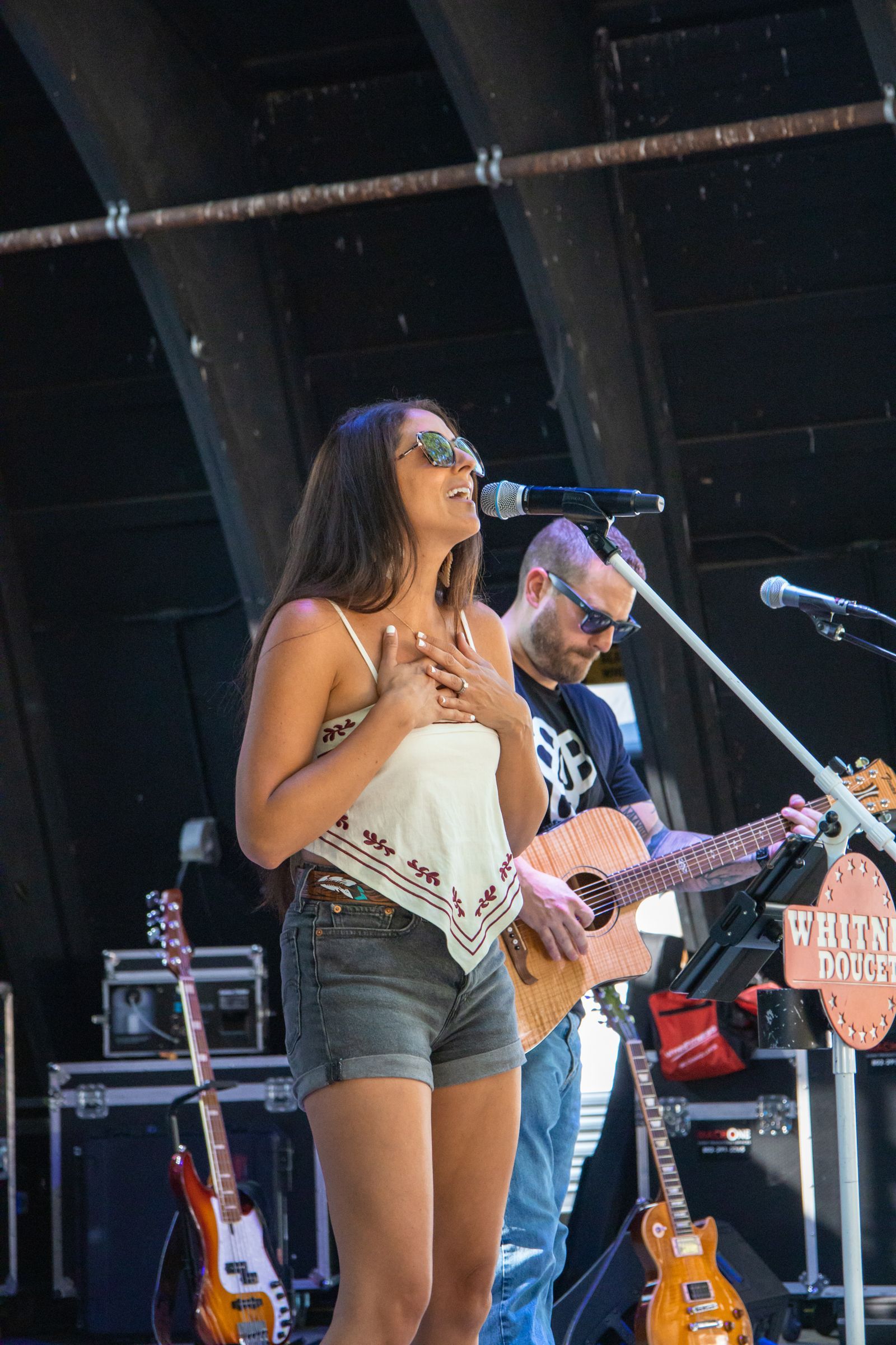 Singer performing onstage with guitarist, holding a microphone and singing outdoors at a live concert