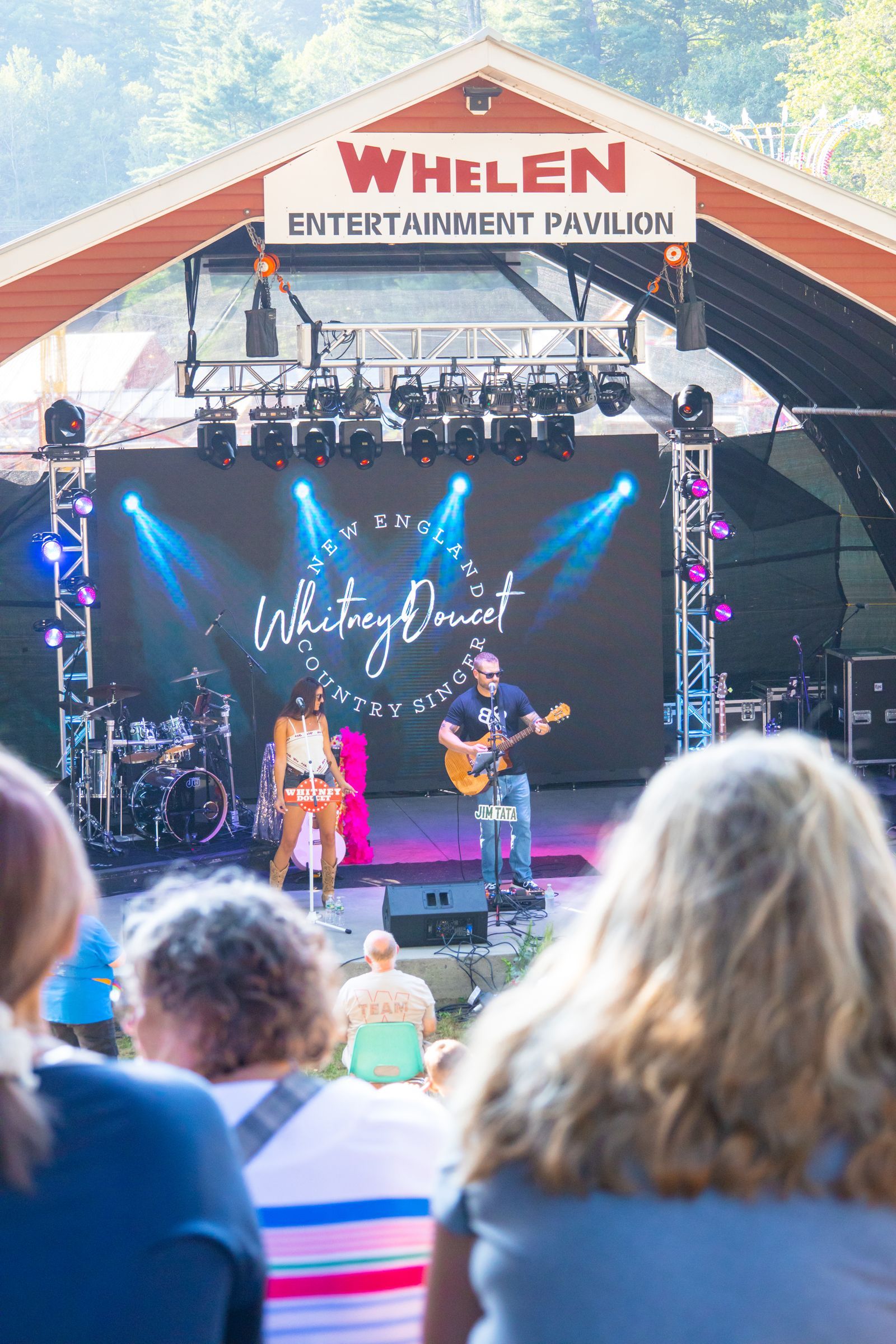 Outdoor concert at Whelen Entertainment Amphitheater, with performers on stage and audience seated in front.