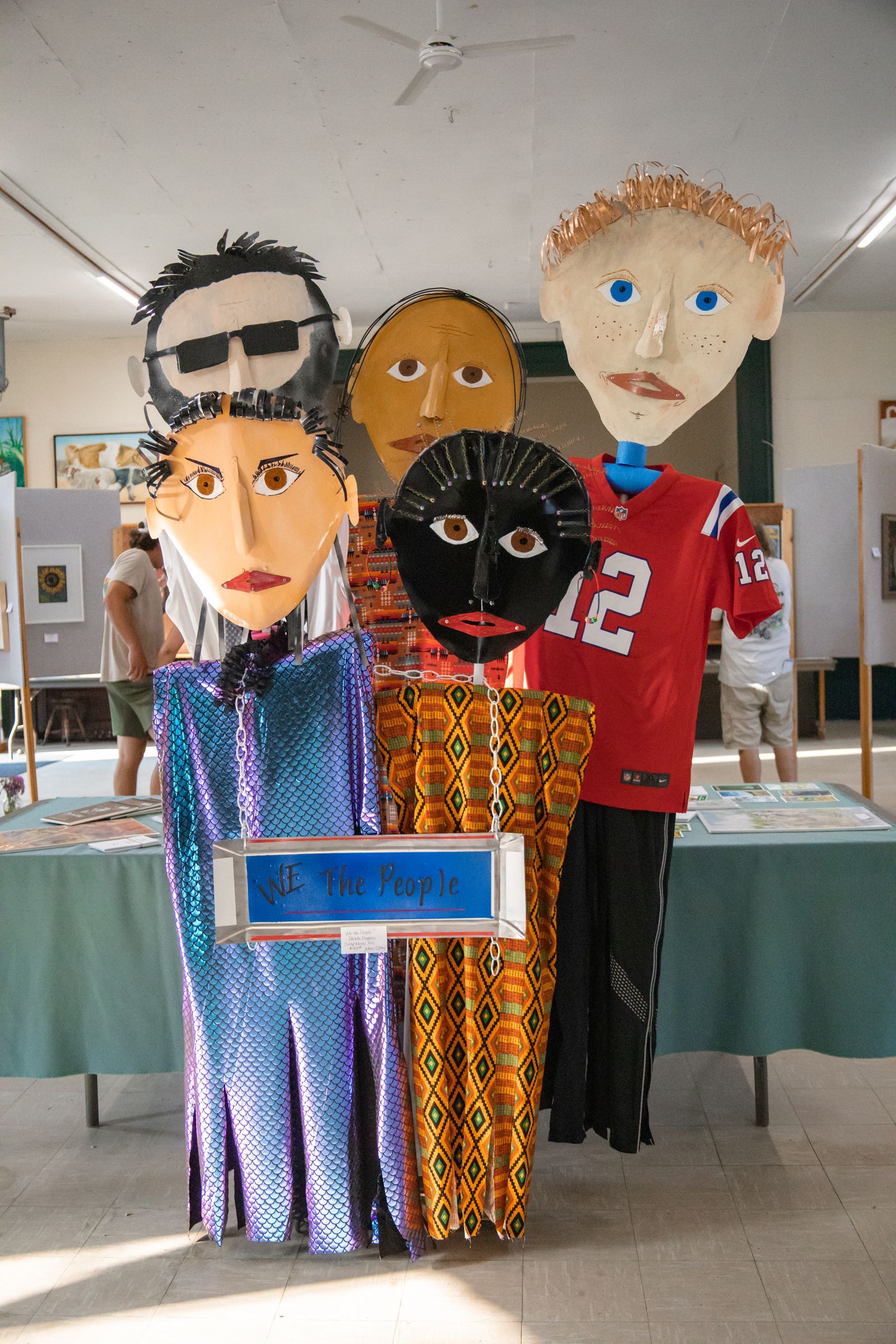 Colorful paper-mâché figures in a display room, with one holding a blue sign and one red jersey figure in front.