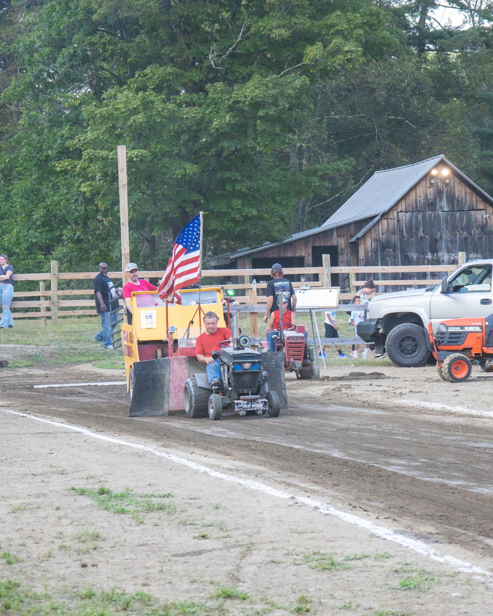 Small tractor pulling a decorated cart with an American flag on a muddy track at a rural fair