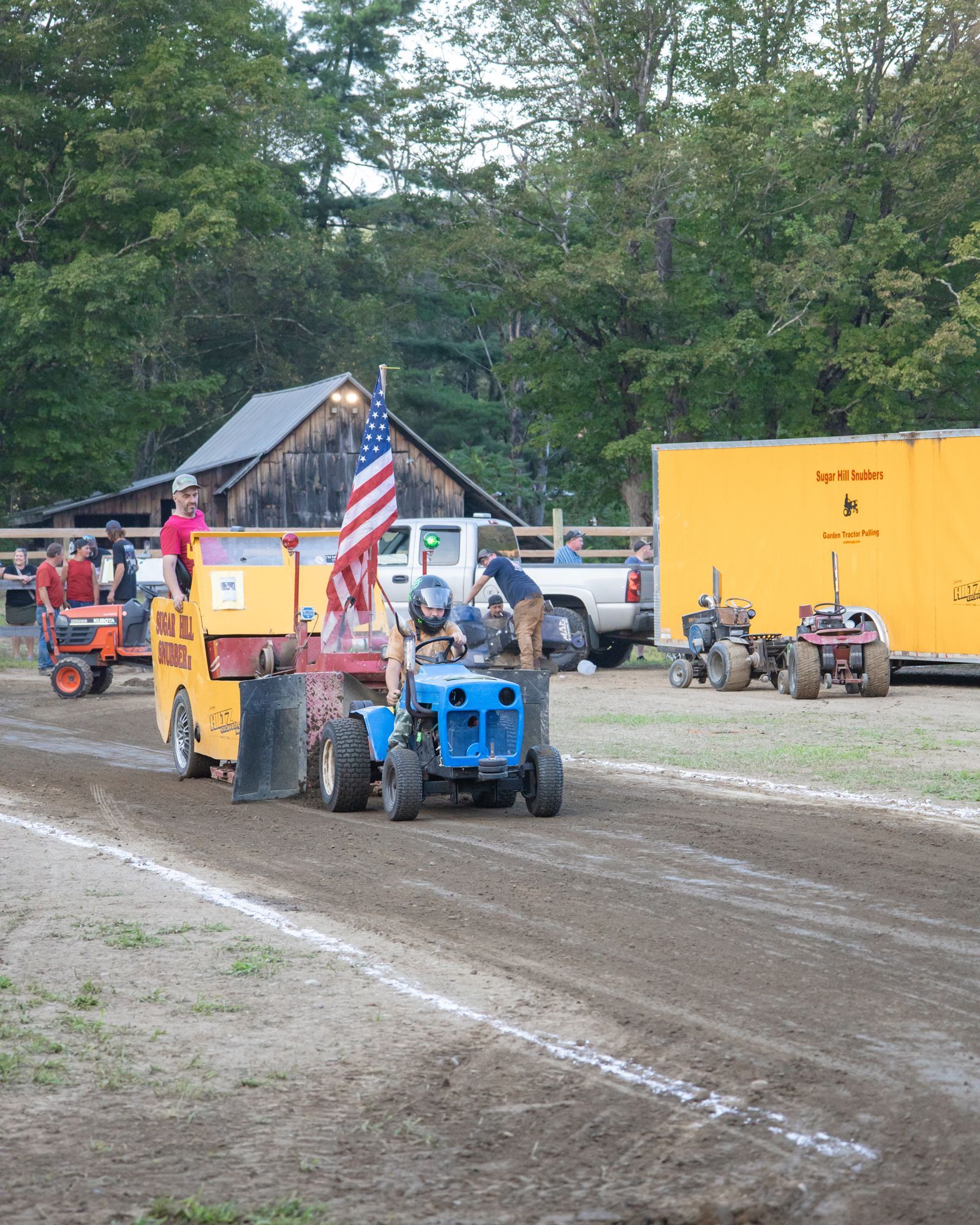 Blue tractor pulls a yellow sled on a muddy dirt track at an outdoor fair, with spectators nearby.