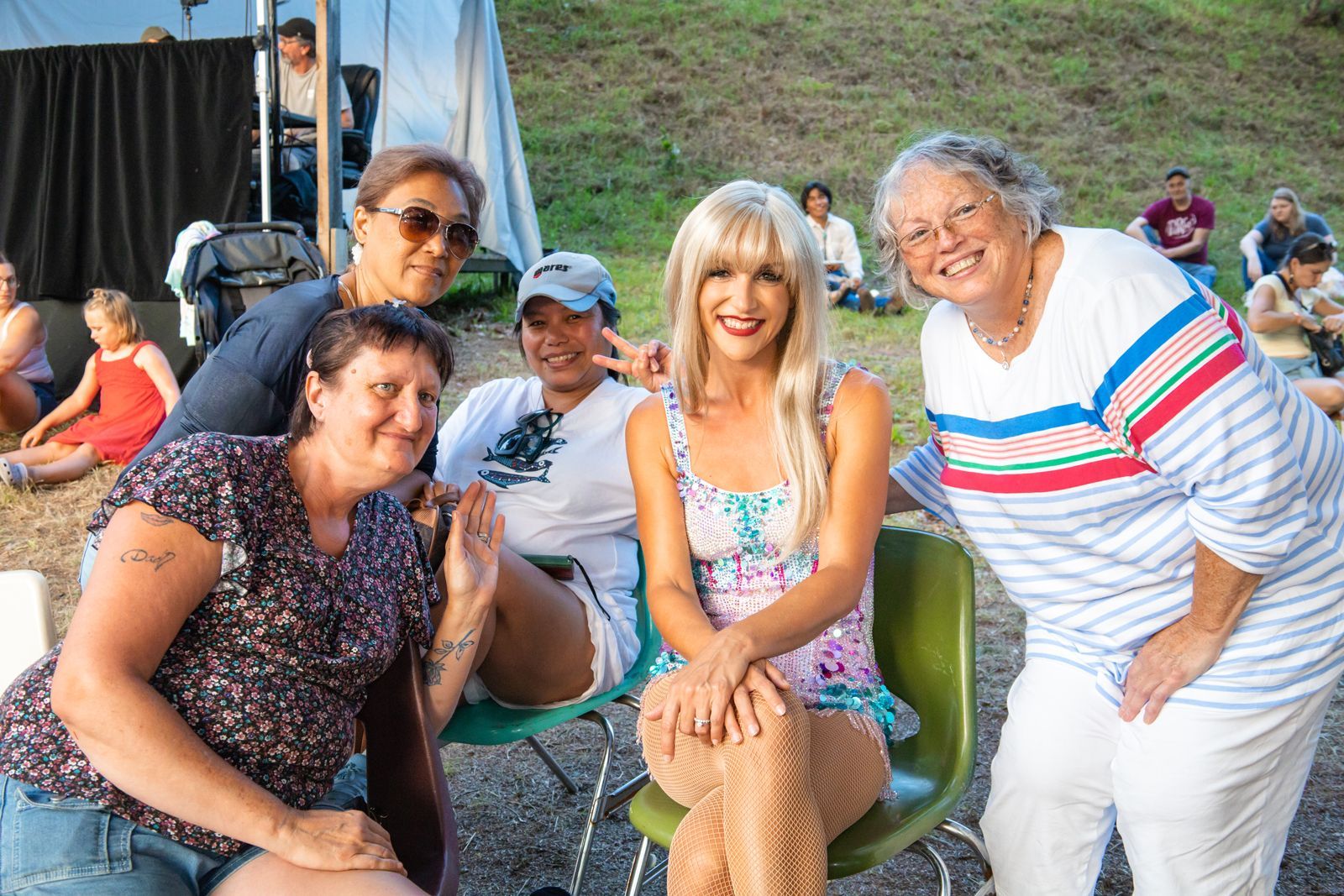 Four friends smiling on outdoor folding chairs at a lively event, with a blonde woman in the center.