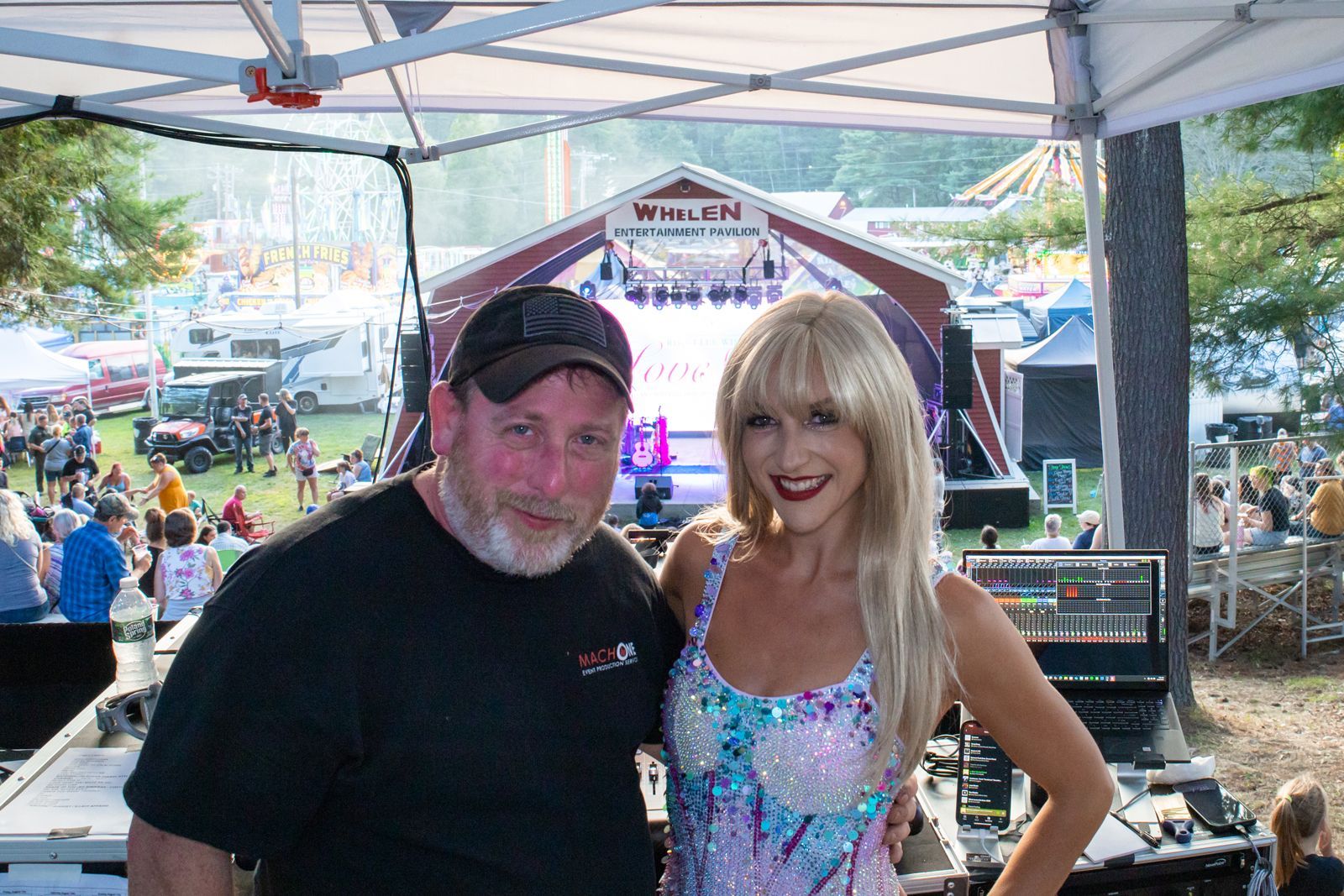 Two people smiling under a canopy at an outdoor event, with stage equipment and a crowd in the background.