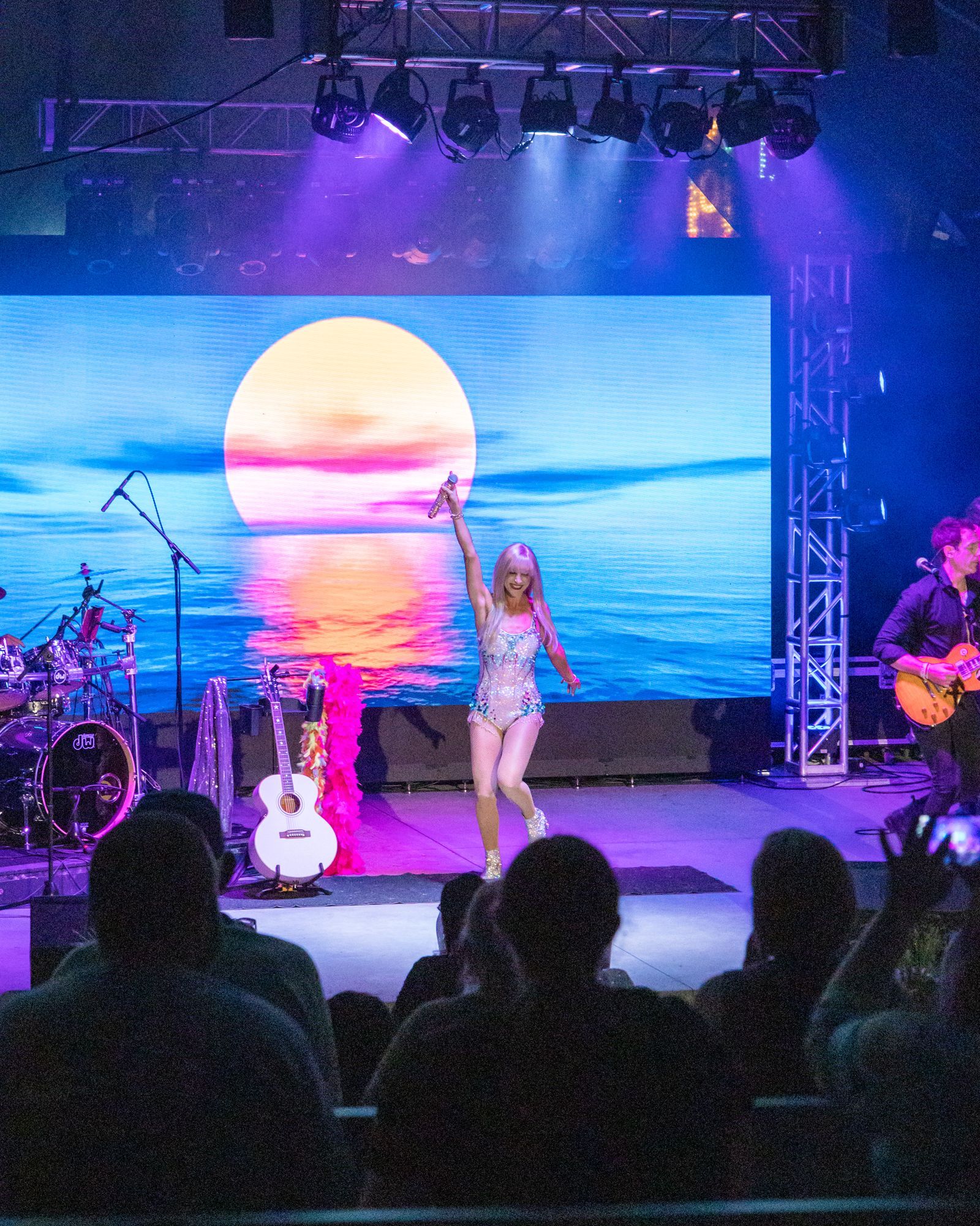 Singer on stage before a sunset ocean backdrop, with guitars and audience silhouettes in front.