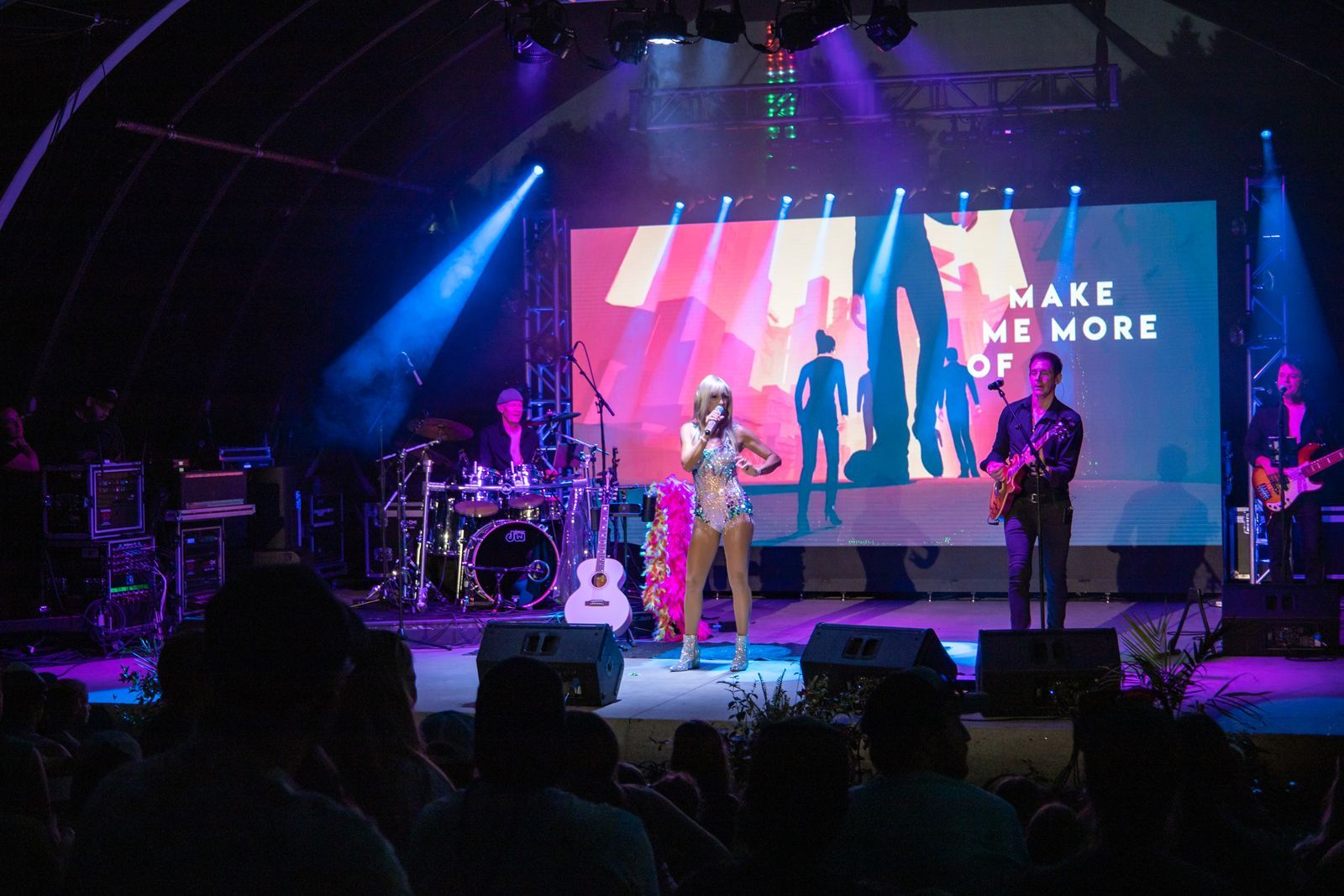 Band performing on a lit concert stage with colorful screens and audience silhouettes in front