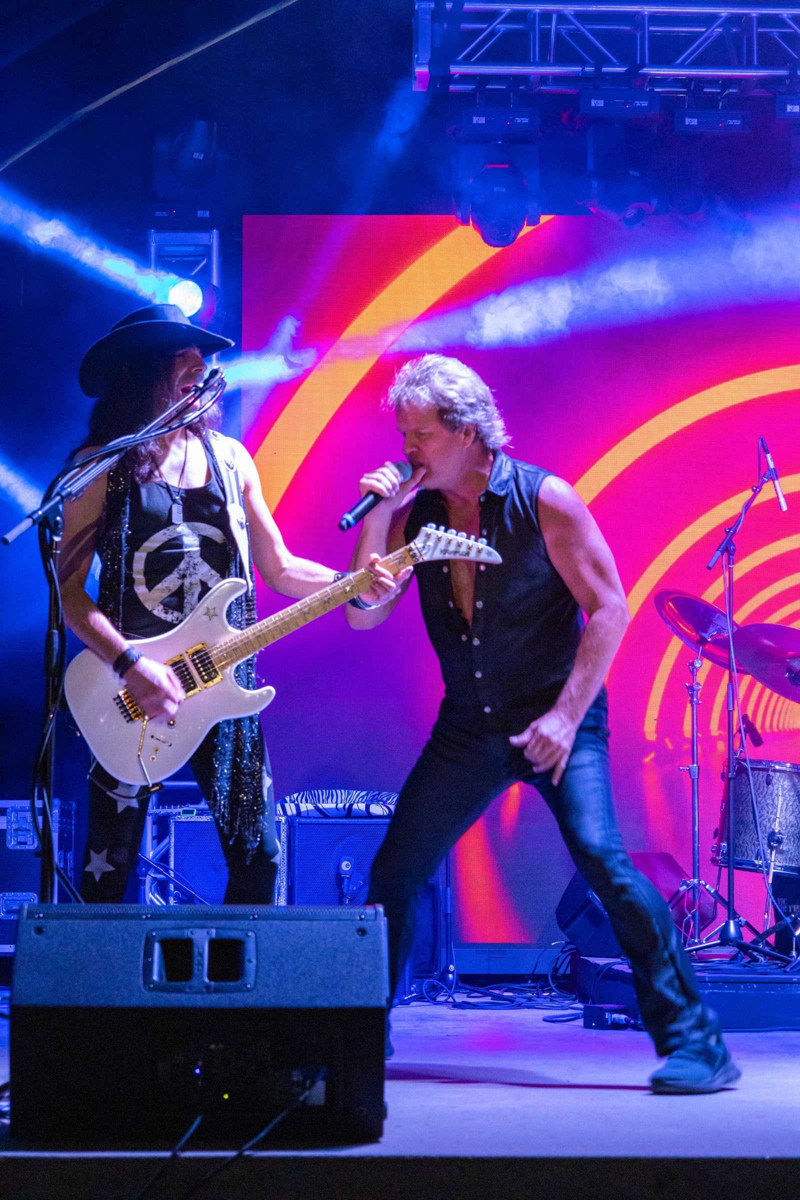 Two musicians performing onstage, one singing into a mic with a guitar against a bright red and blue backdrop
