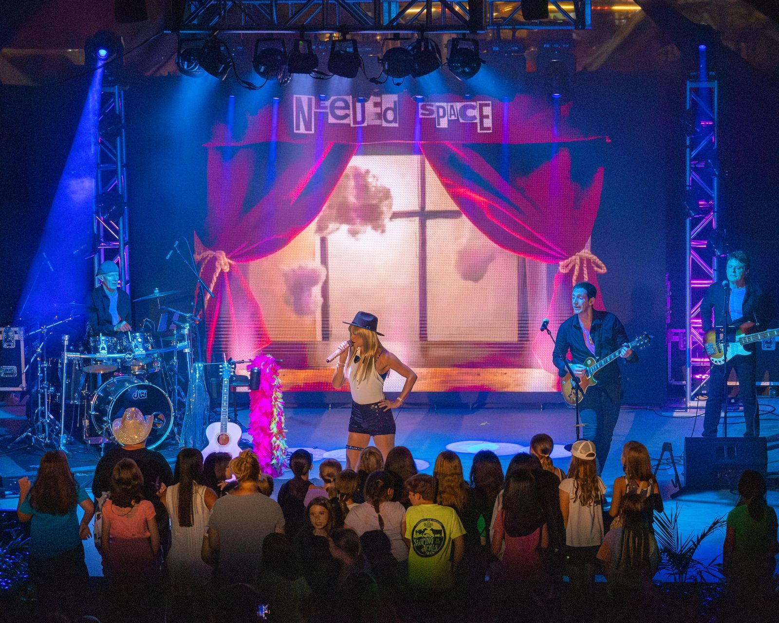 Live rock band performing onstage under colorful lights, with an audience watching from the front.