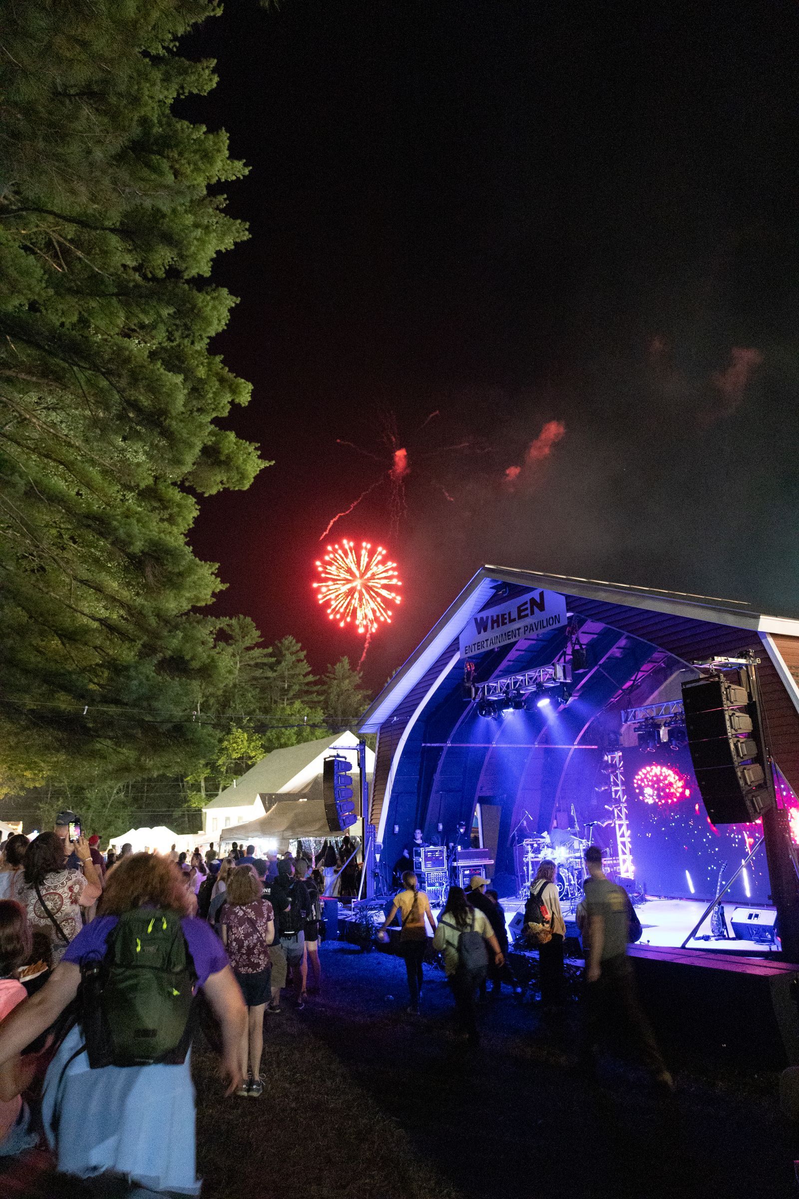 Night concert crowd by a lit stage with fireworks in the sky and trees at left