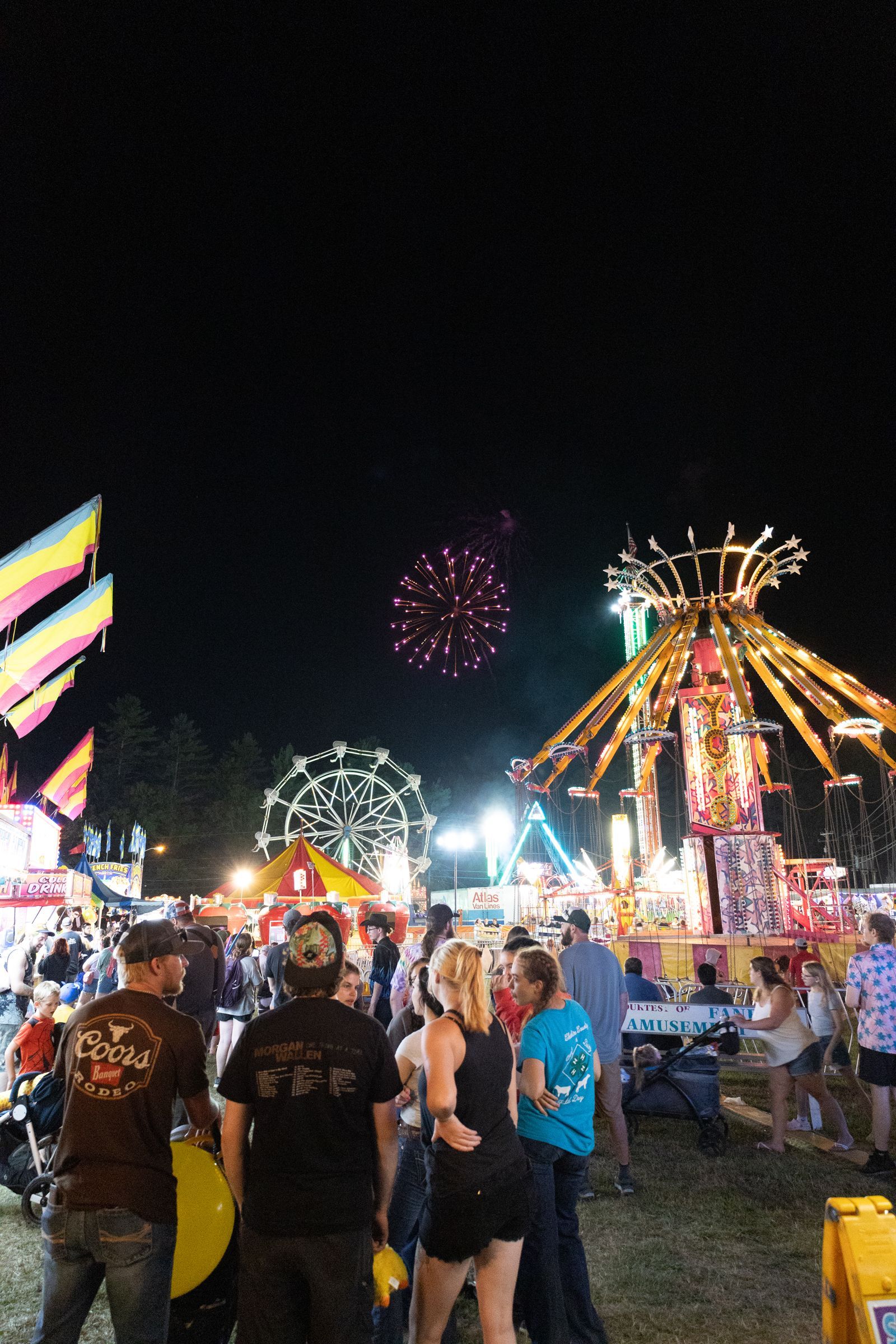 Crowded nighttime fair with carnival rides, bright lights, fireworks, and people standing in the foreground