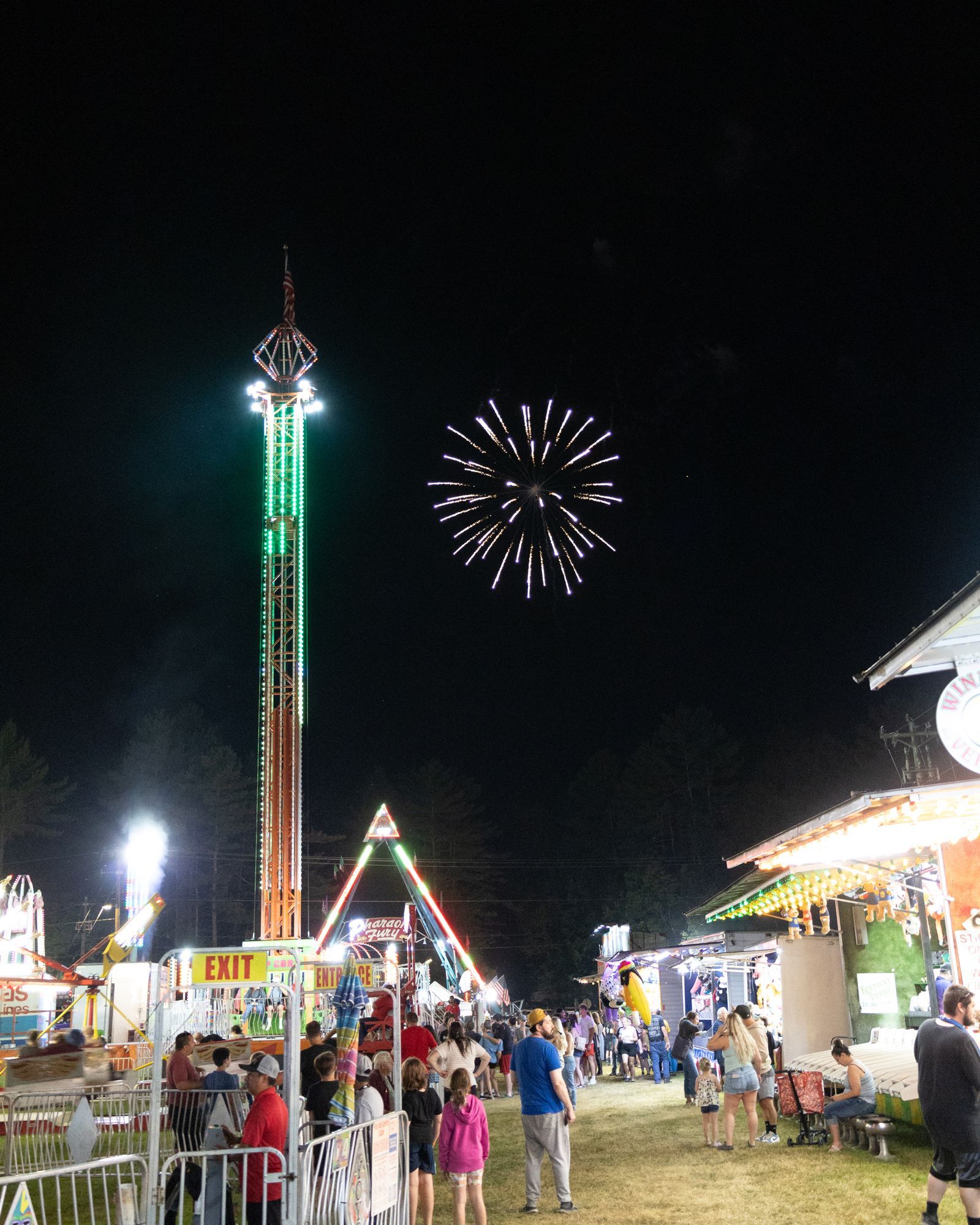 Night fairground with bright rides, crowds, and a fireworks burst in the dark sky
