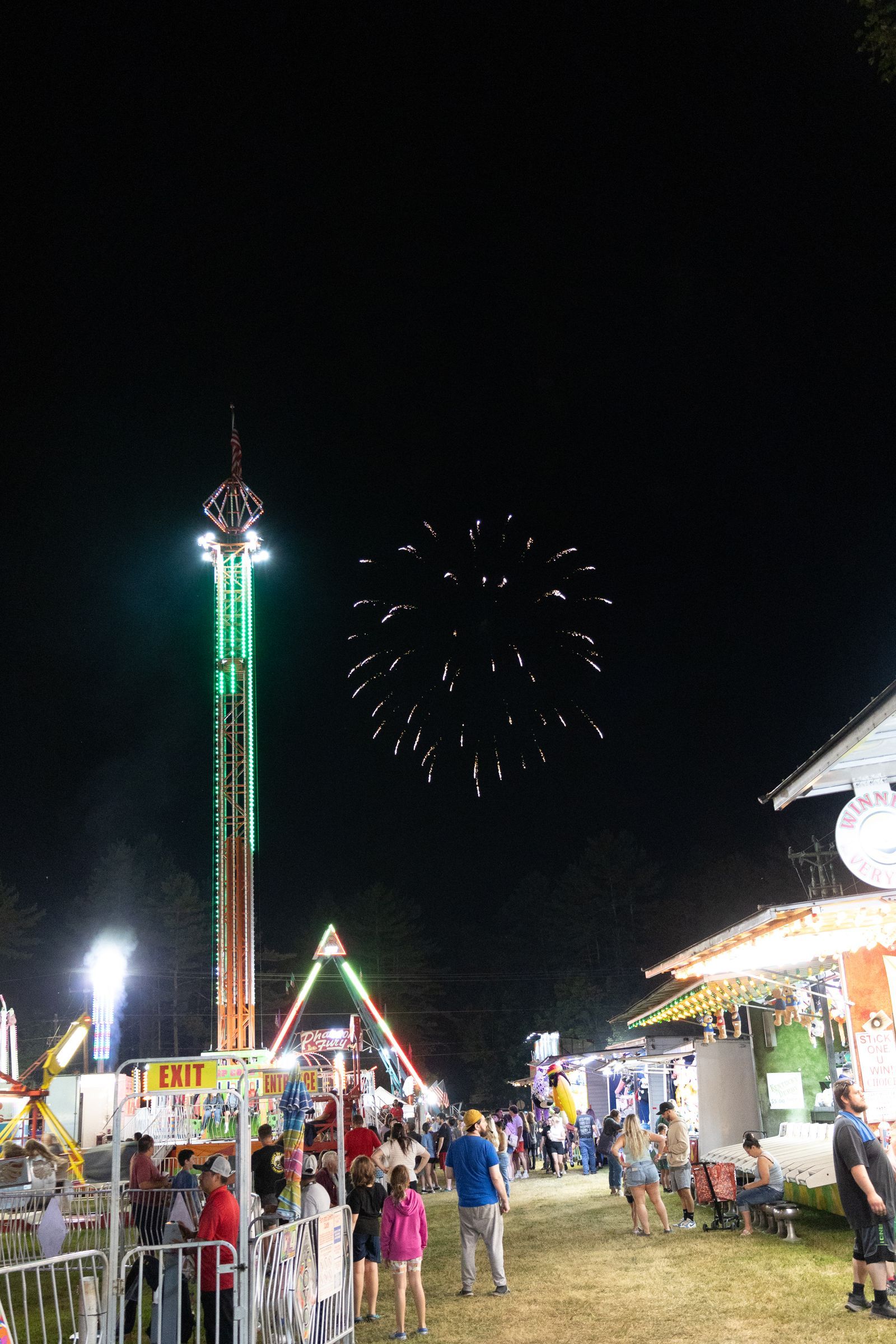 Nighttime carnival with a tall lit ride, fireworks overhead, and a crowd near bright stalls