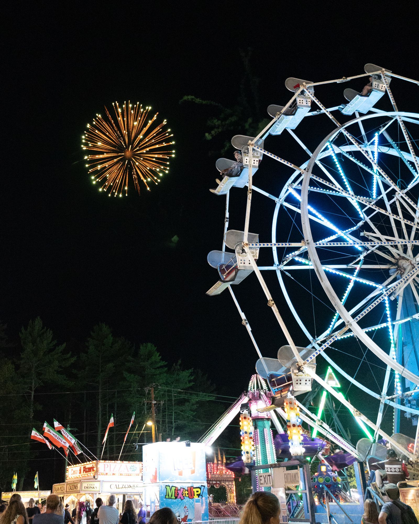 Night carnival with a lit Ferris wheel, fireworks, and colorful rides against a dark sky