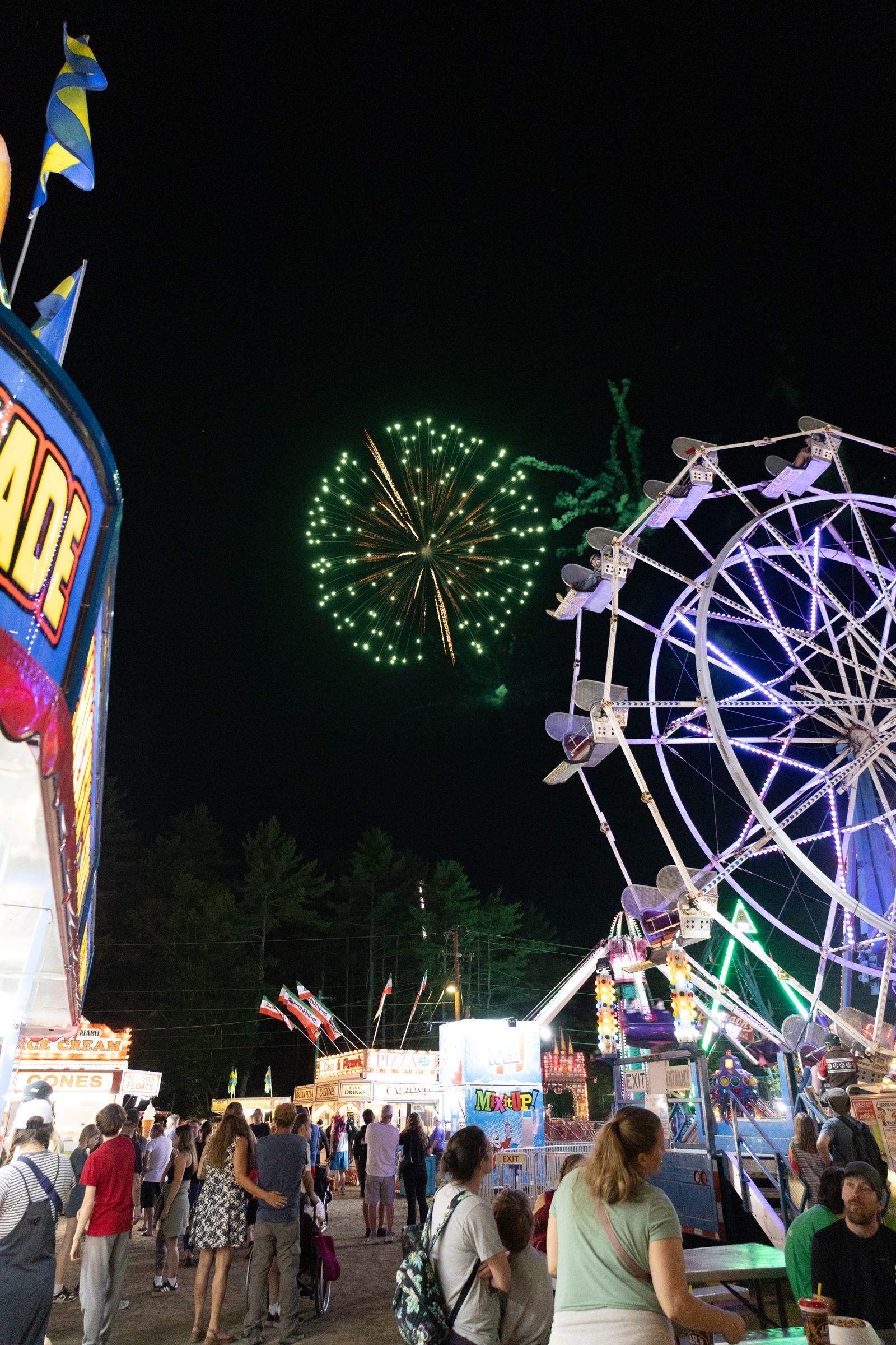 Crowded fairground at night with a Ferris wheel and fireworks in the sky