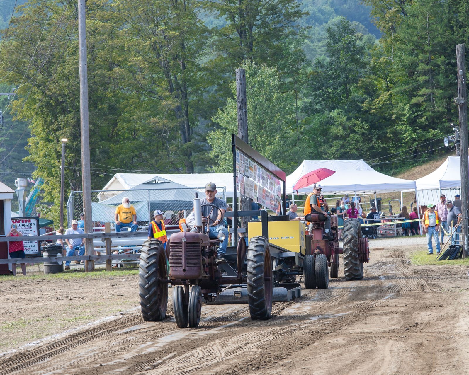 Tractors lined up on a muddy track at an outdoor fair with tents and trees in the background