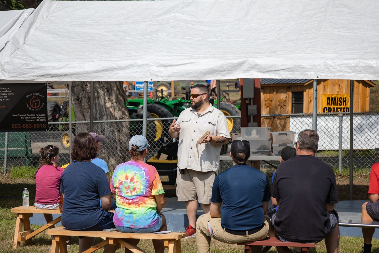 Man speaking under white tent to seated audience outdoors at a community event