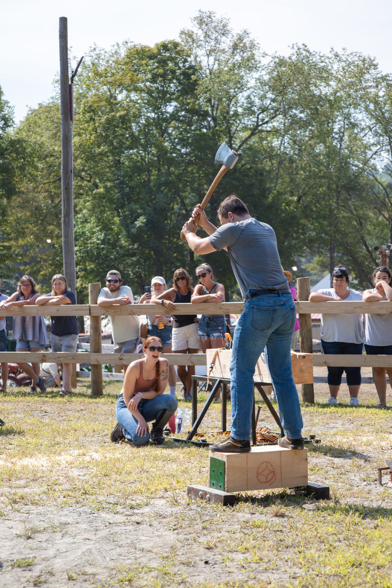 Person swings an axe at a target block outdoors while a crowd watches behind a wooden barrier.