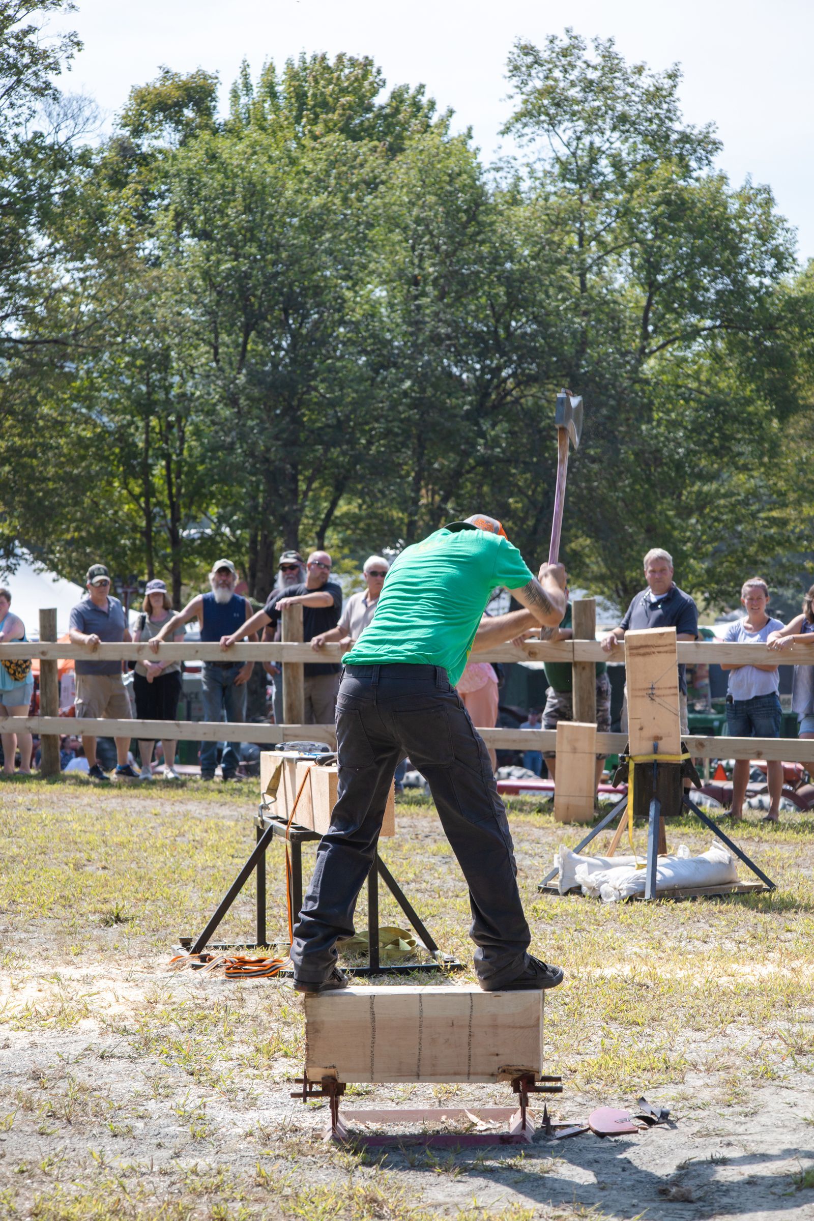 Person in green shirt chopping wood outdoors at a crowd event.