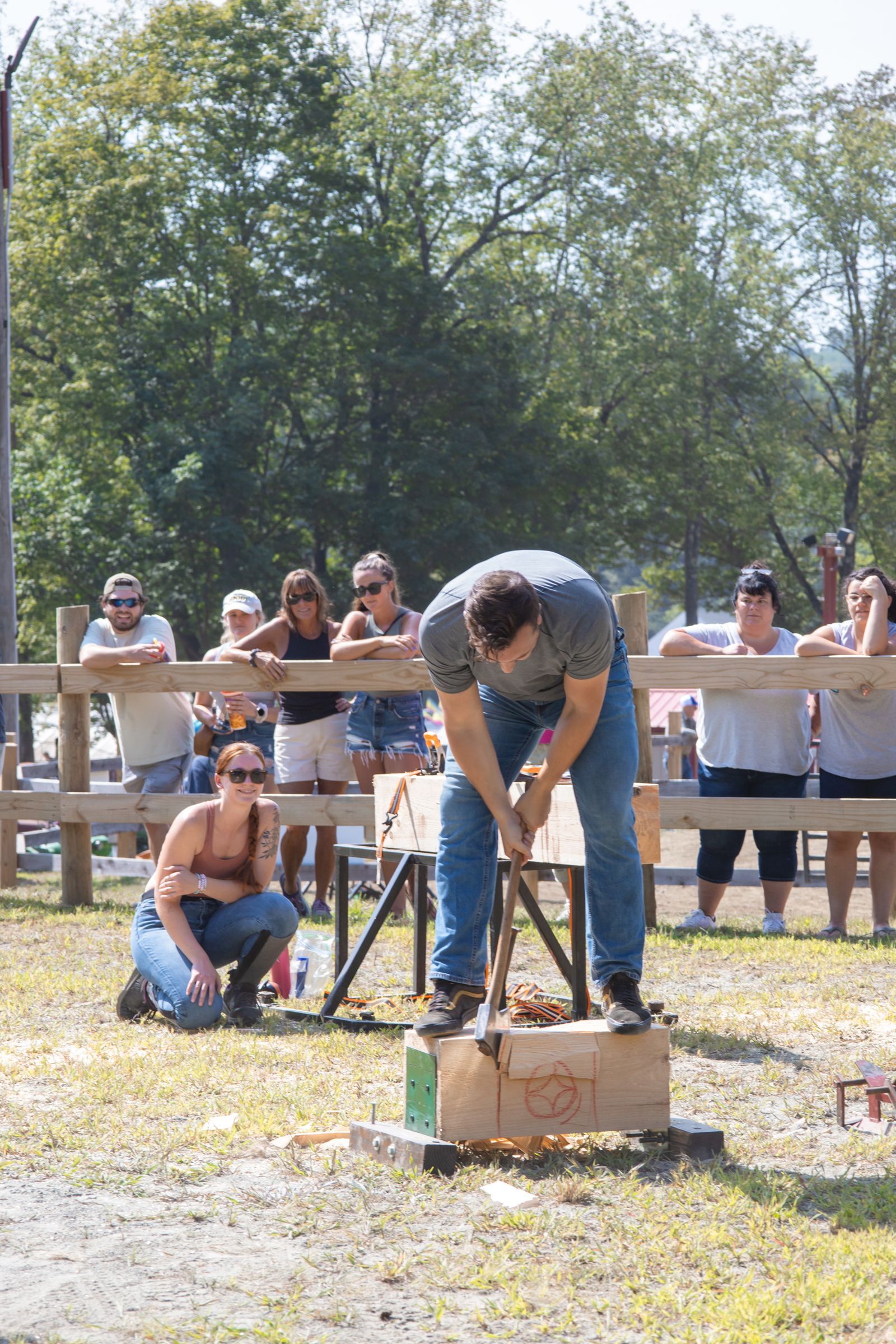 Person bent over swinging a sledgehammer at a block outdoors, with spectators behind a fence.