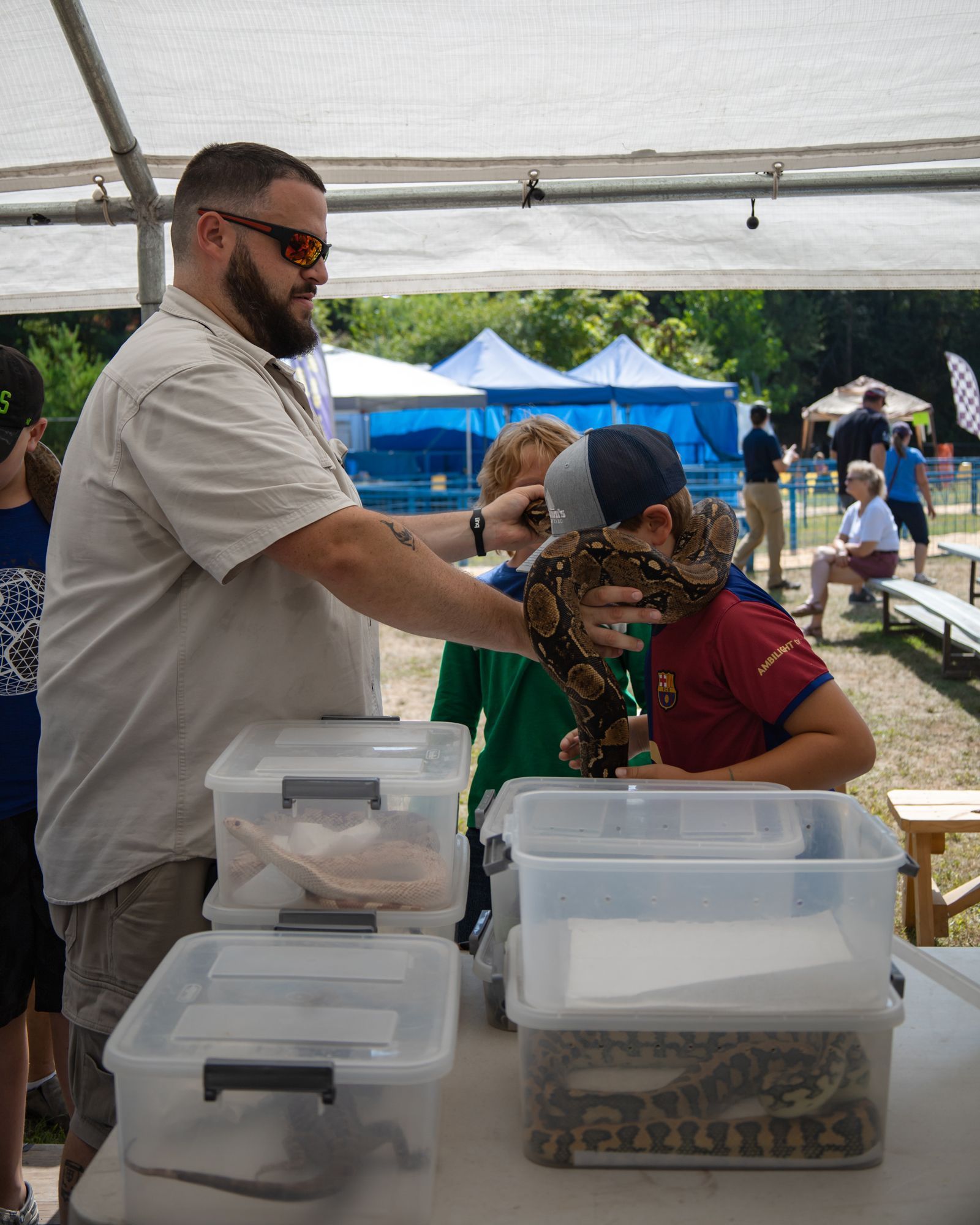 Person handling a snake at an outdoor booth, with clear plastic tubs in the foreground and tents behind.