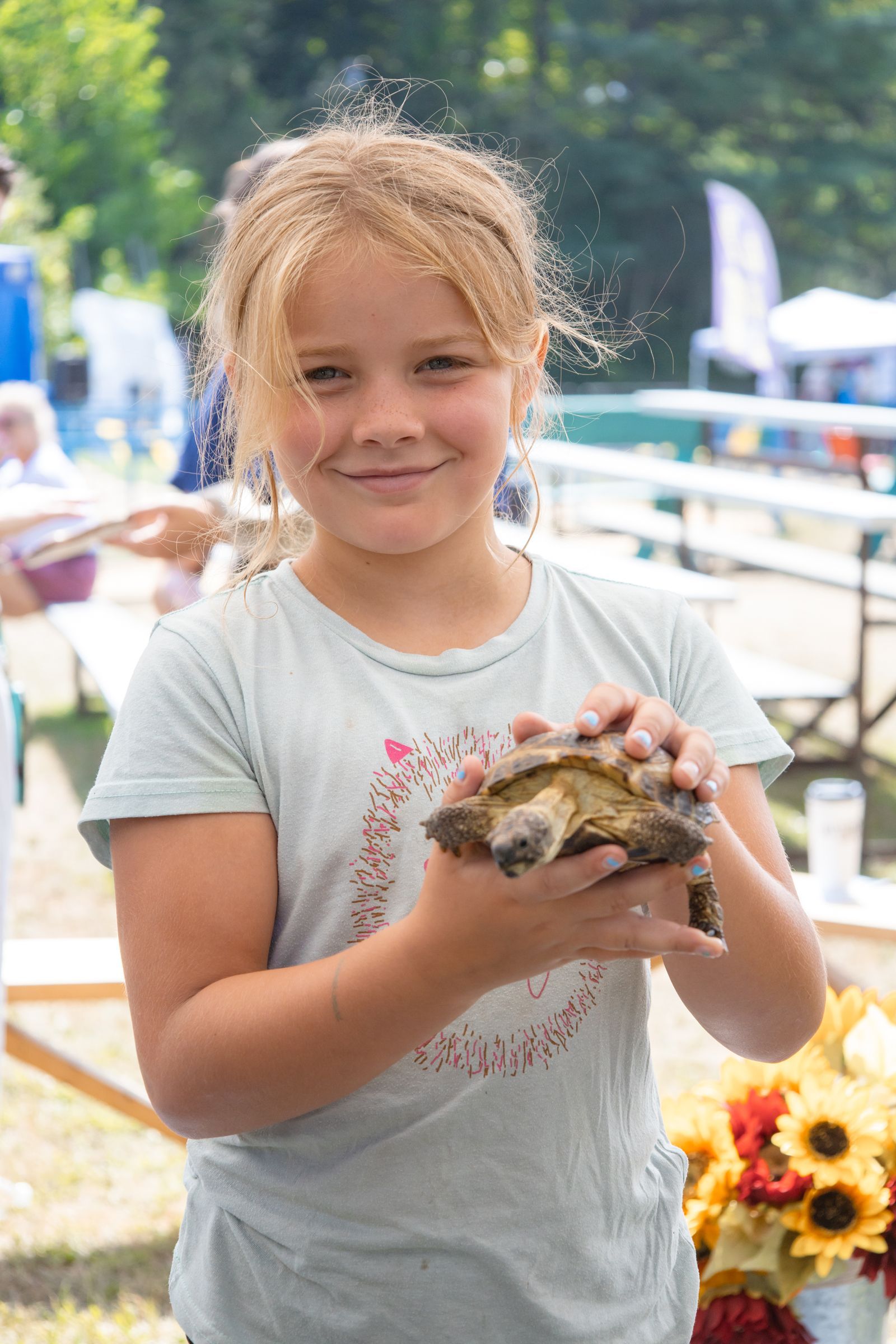 Smiling child holding a small turtle at an outdoor event, with flowers in the background