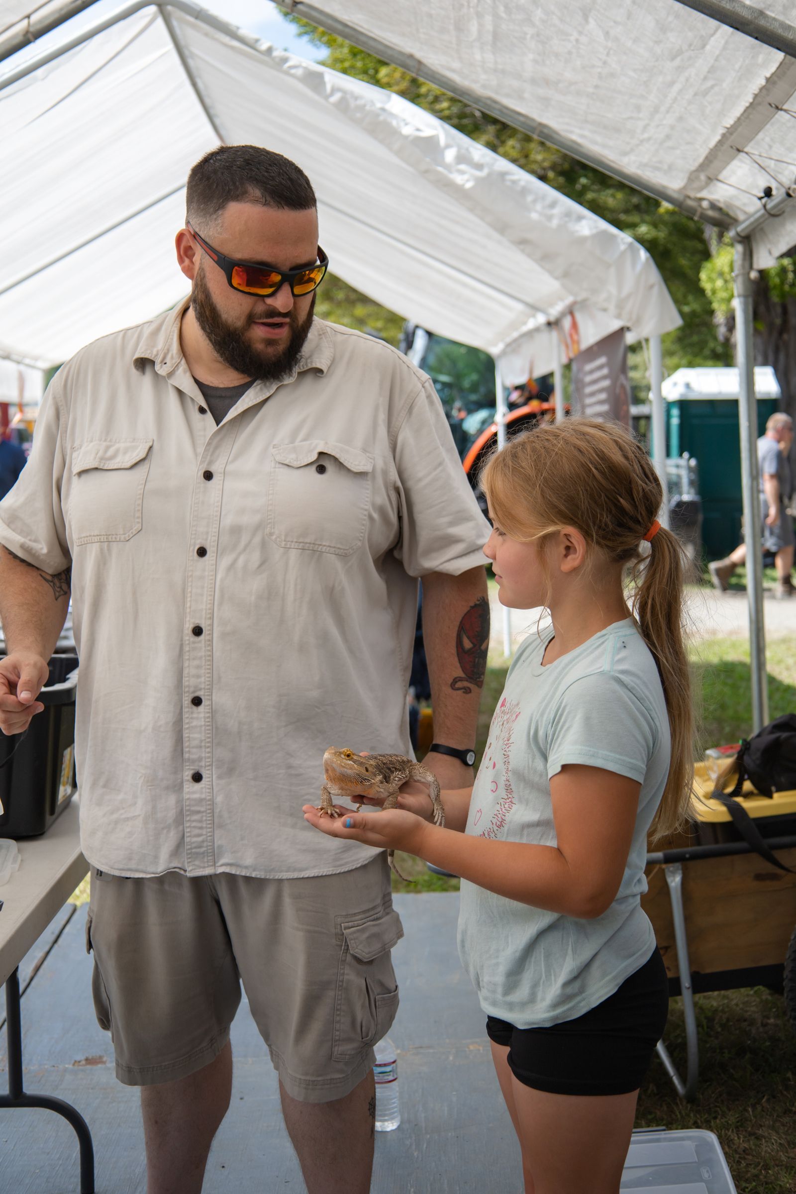 Man in beige shirt speaking to girl holding a tray inside a white tent