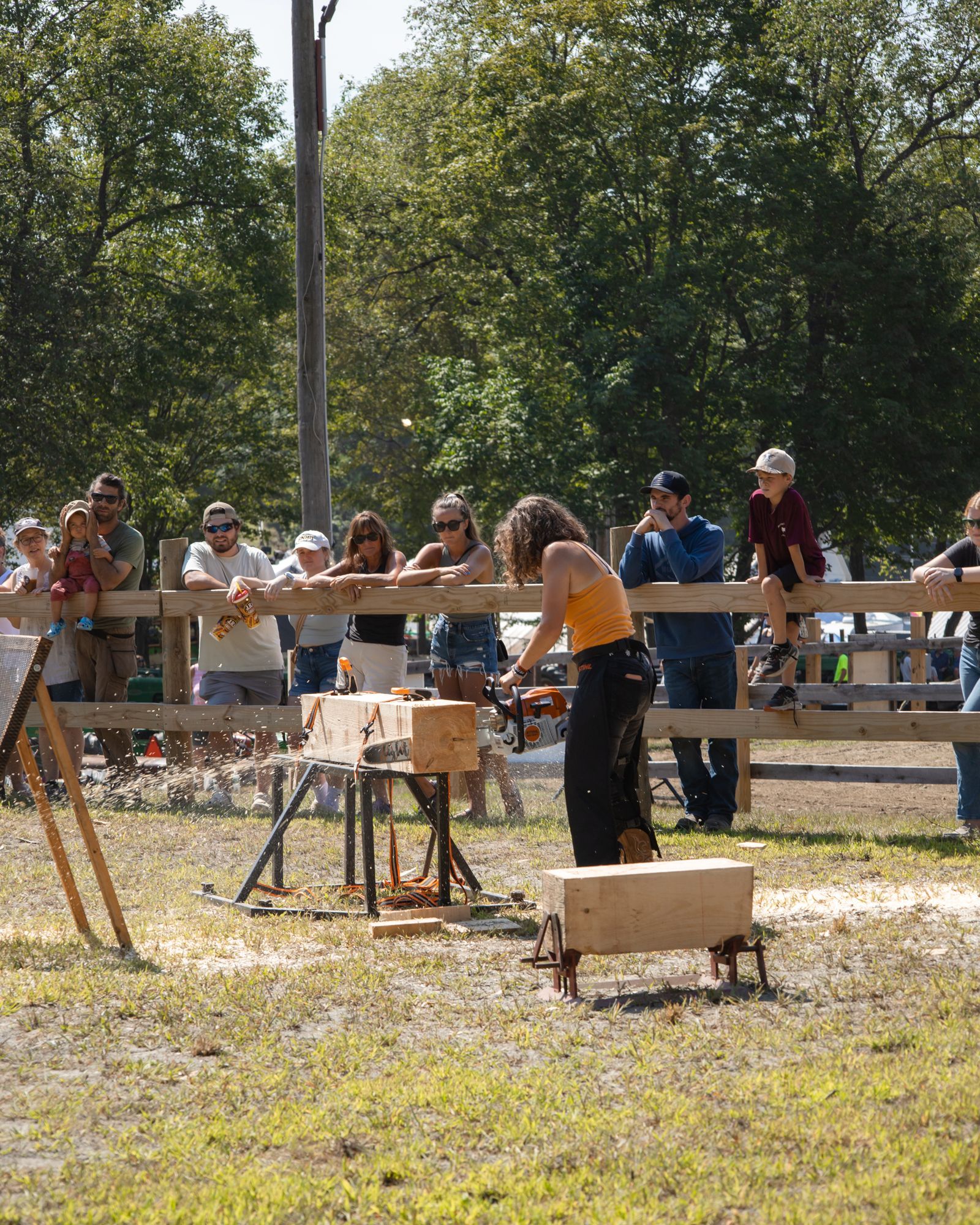 Person sawing wood outdoors while a small crowd watches behind a fence.