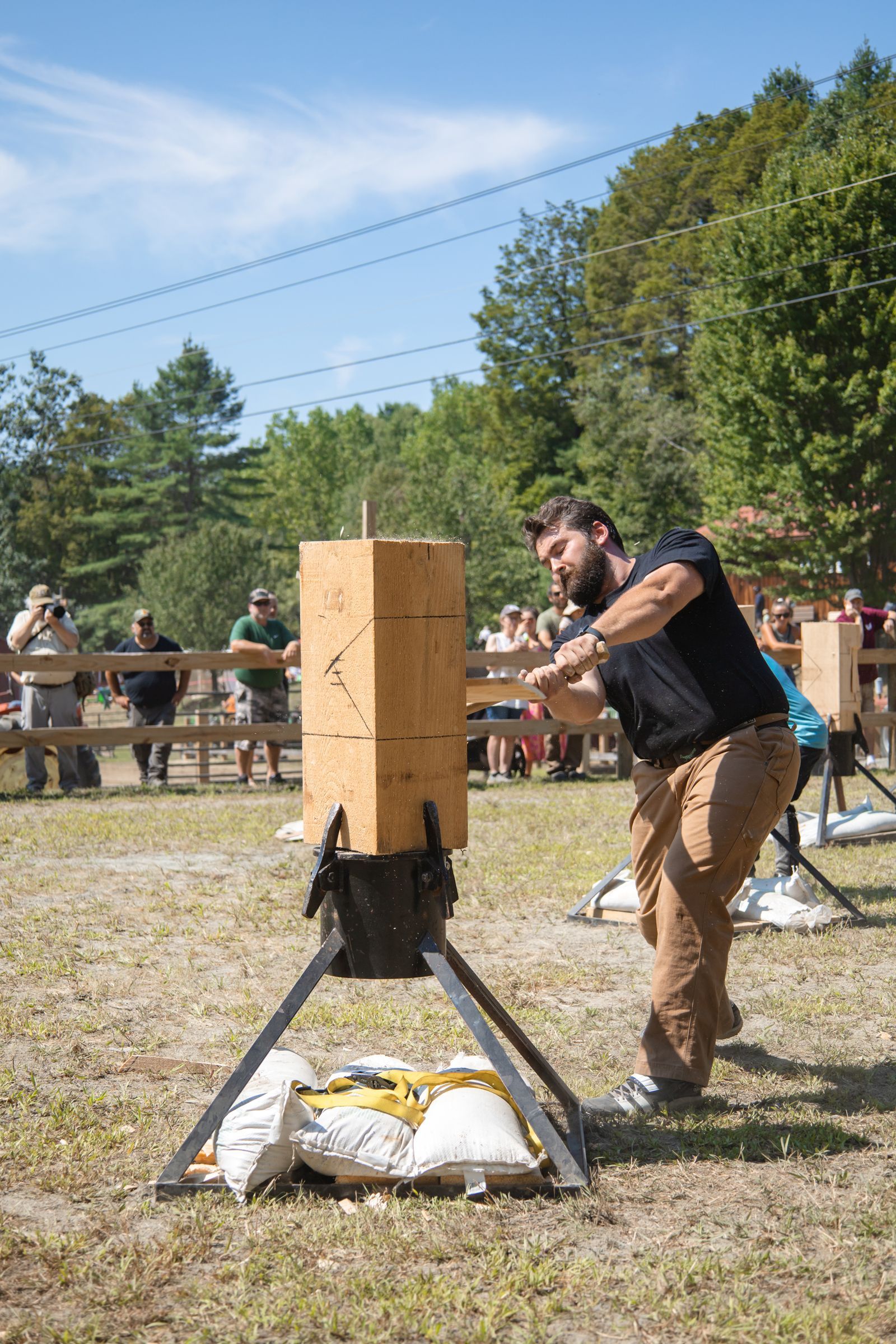 Man striking a wooden target with a mallet at an outdoor event, with spectators and trees in the background