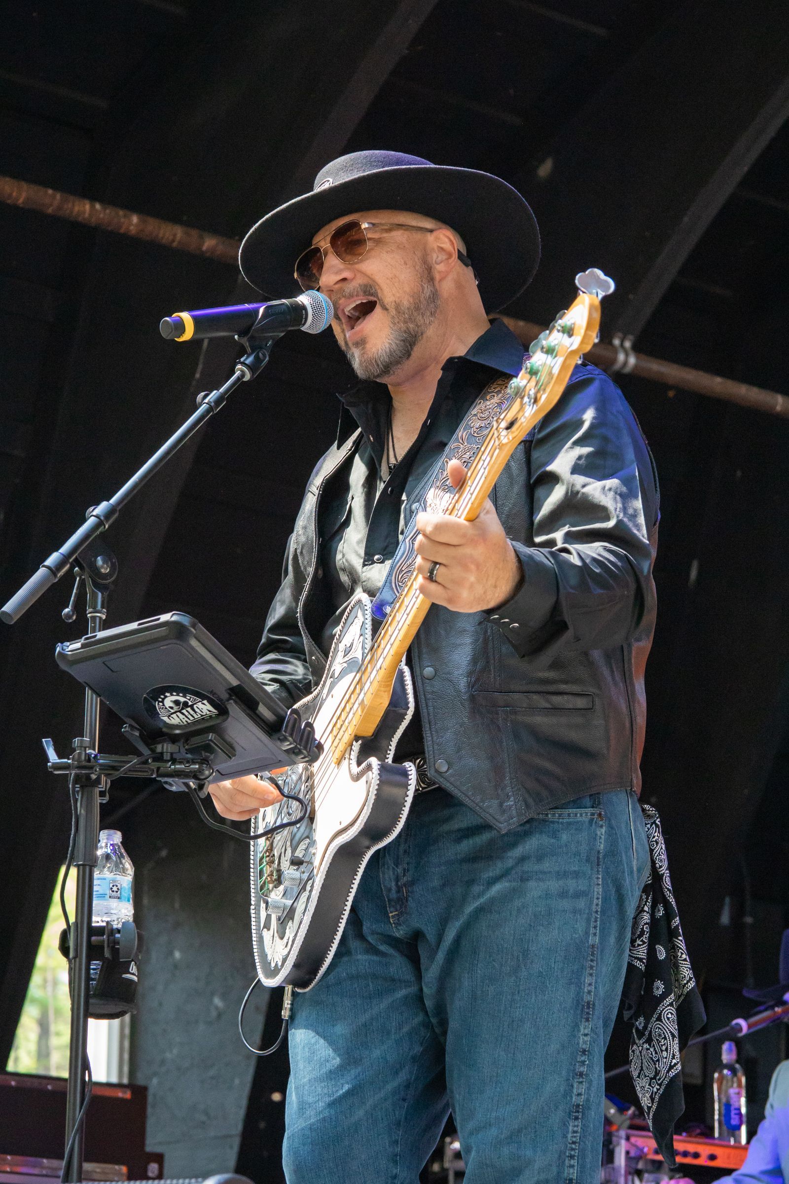 Musician in black hat and jacket sings and plays bass guitar onstage at a microphone