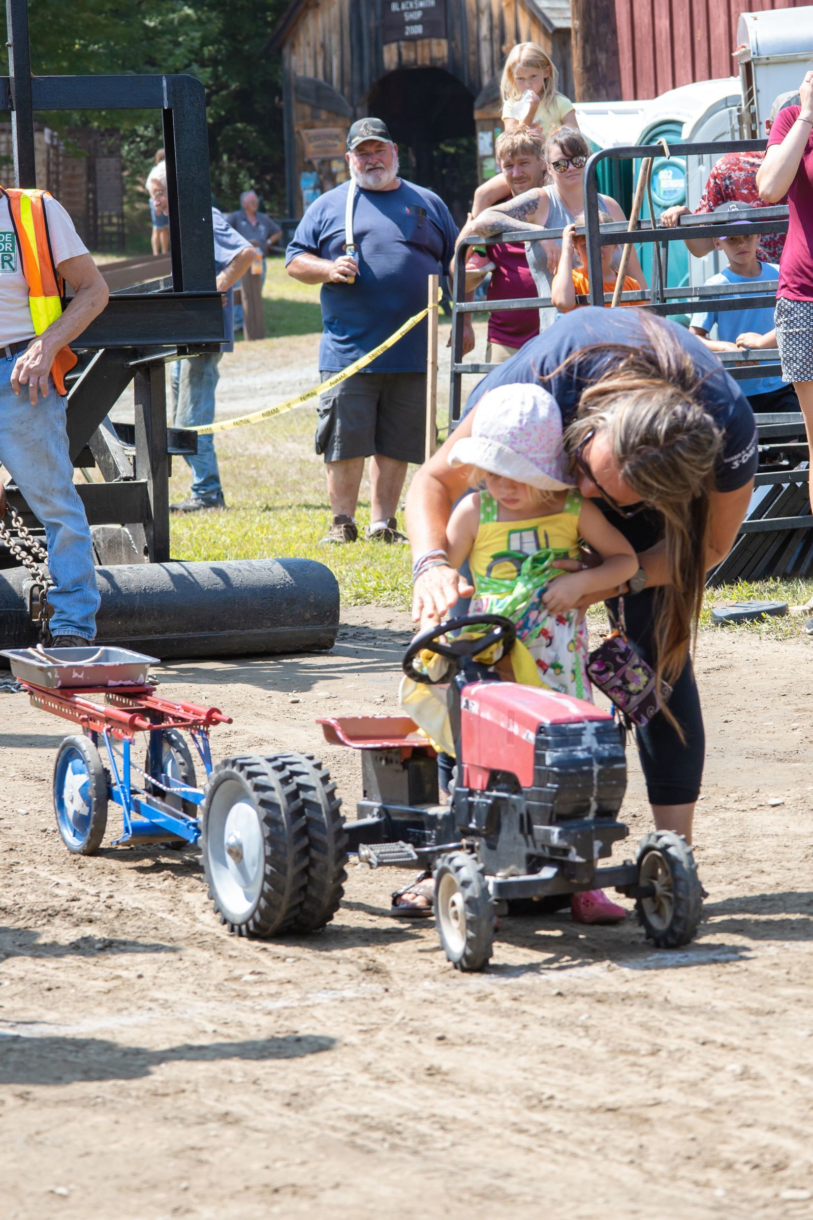 Child leaning into a small red toy car at an outdoor event, with a crowd in the background