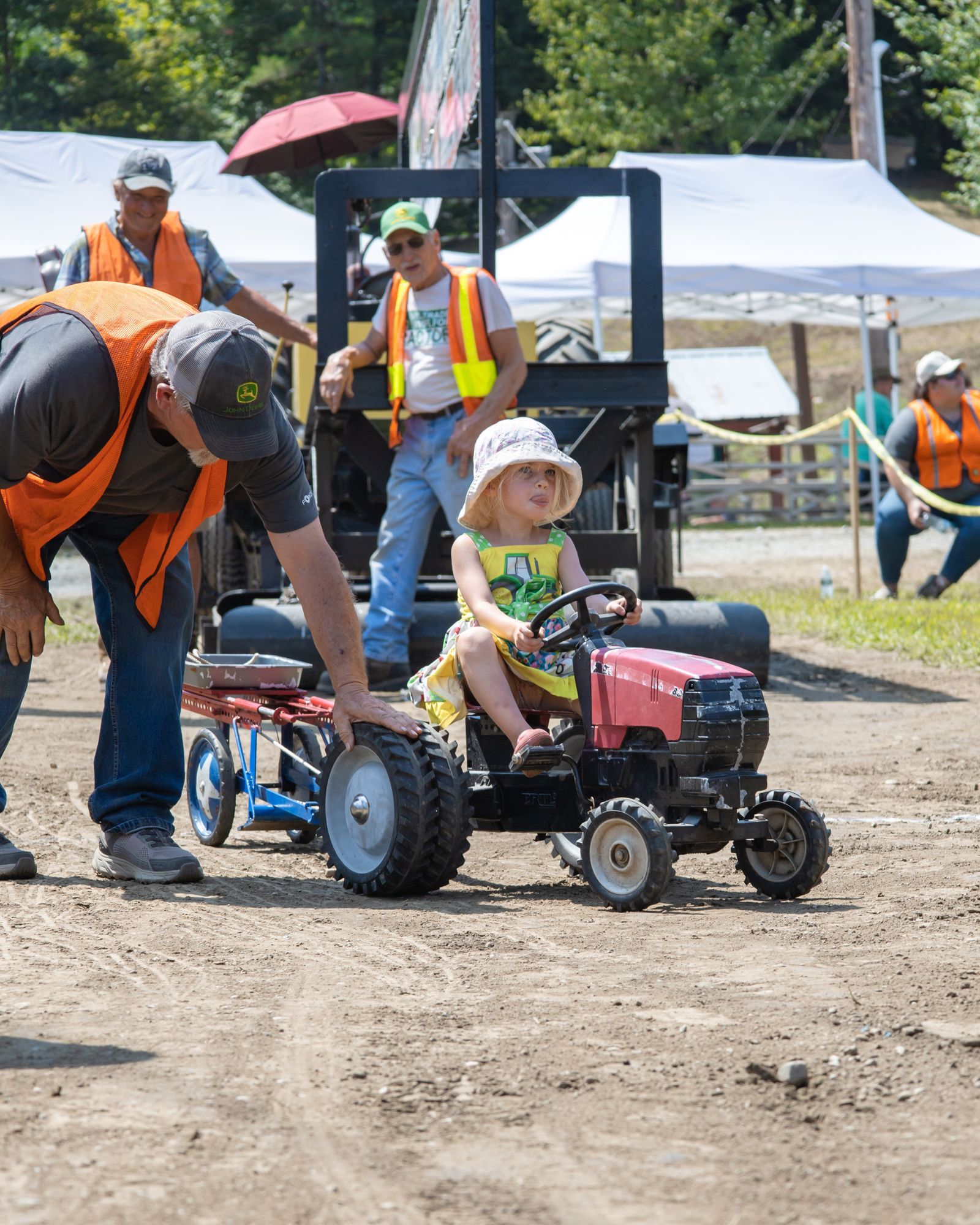 Child driving a small red tractor while adults in safety vests stand nearby at an outdoor event