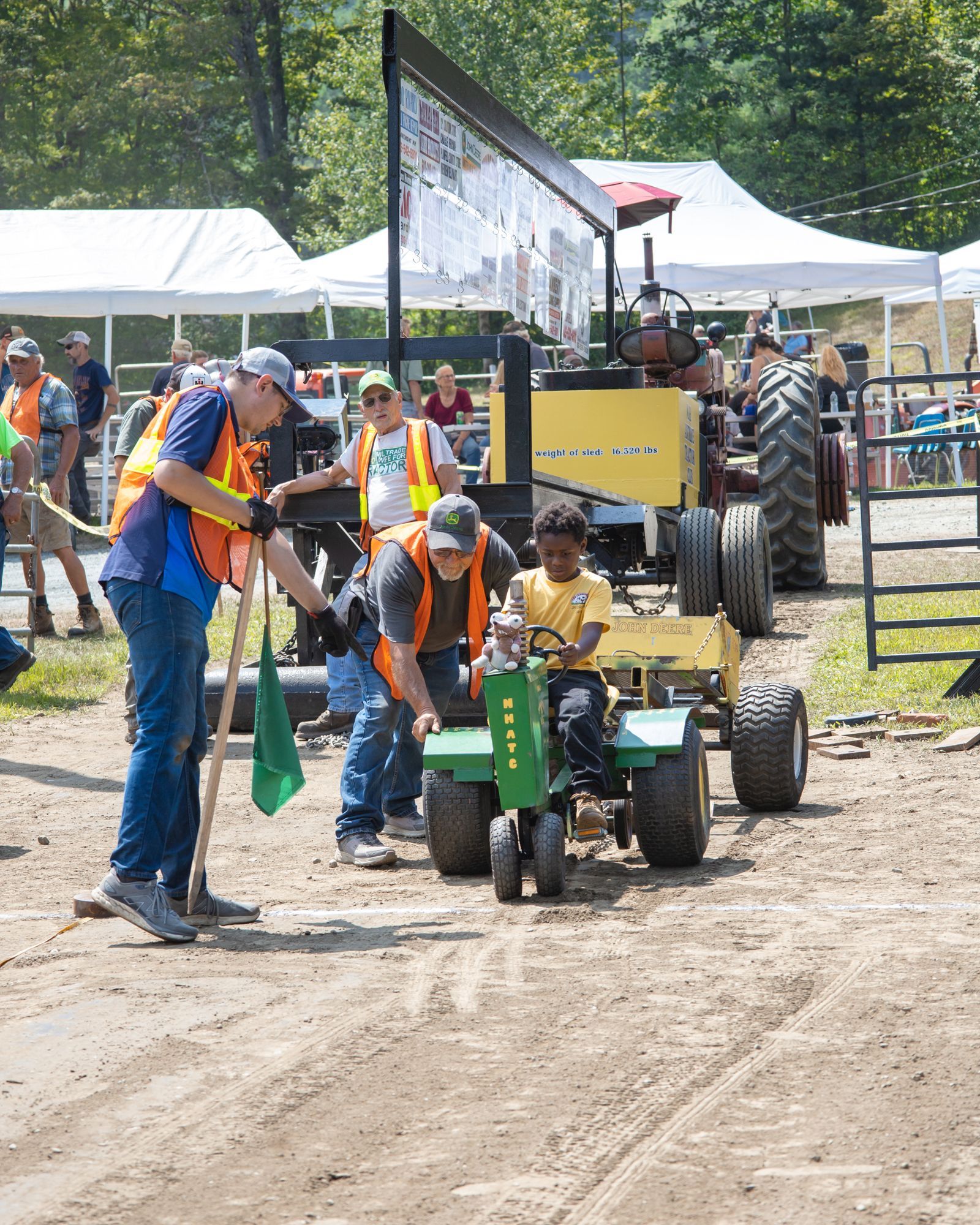 Children on small tractors at a dirt track, guided by adults near tents and a crowd.