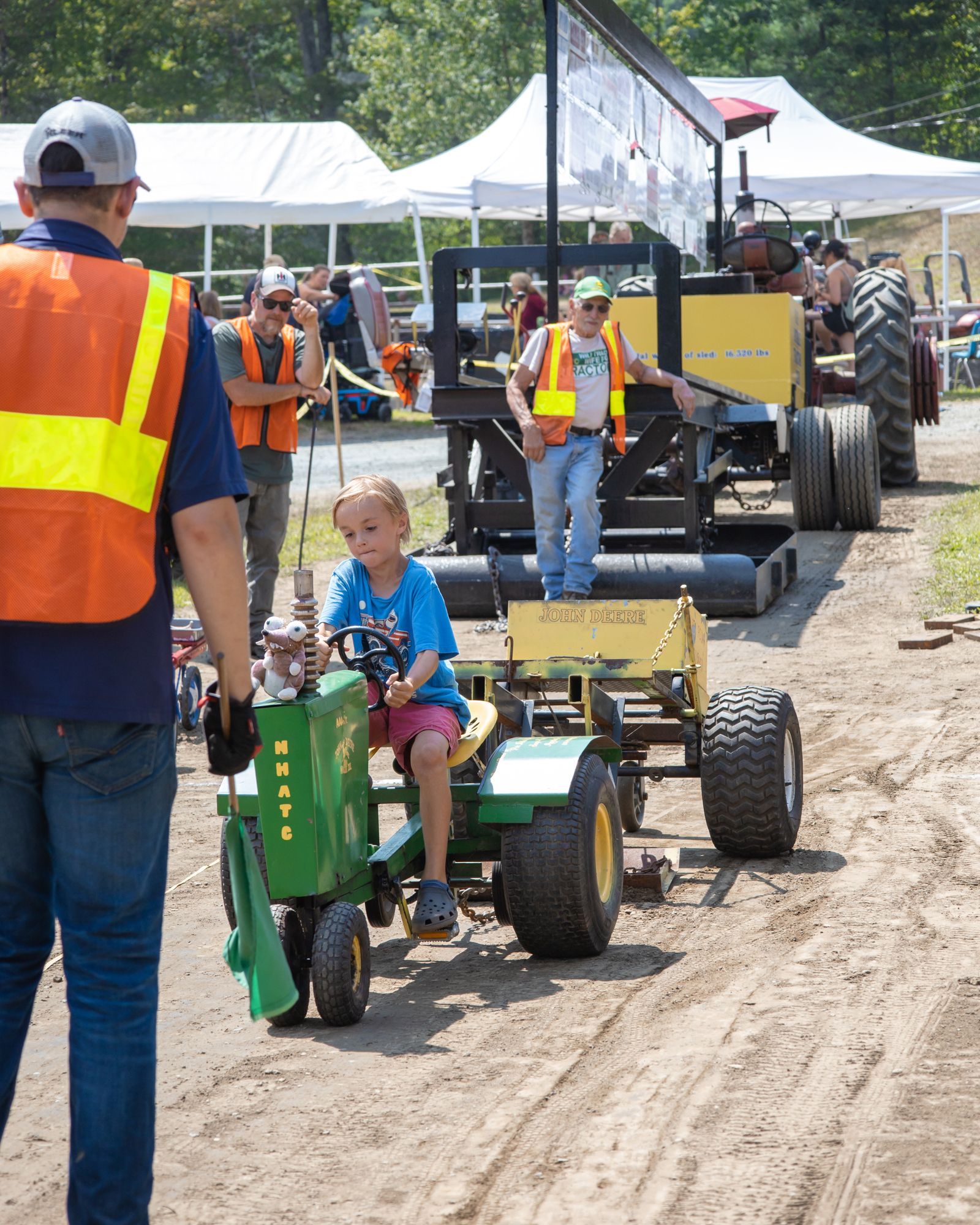 Child driving a green toy tractor at an outdoor event, with adults and other tractors in the background