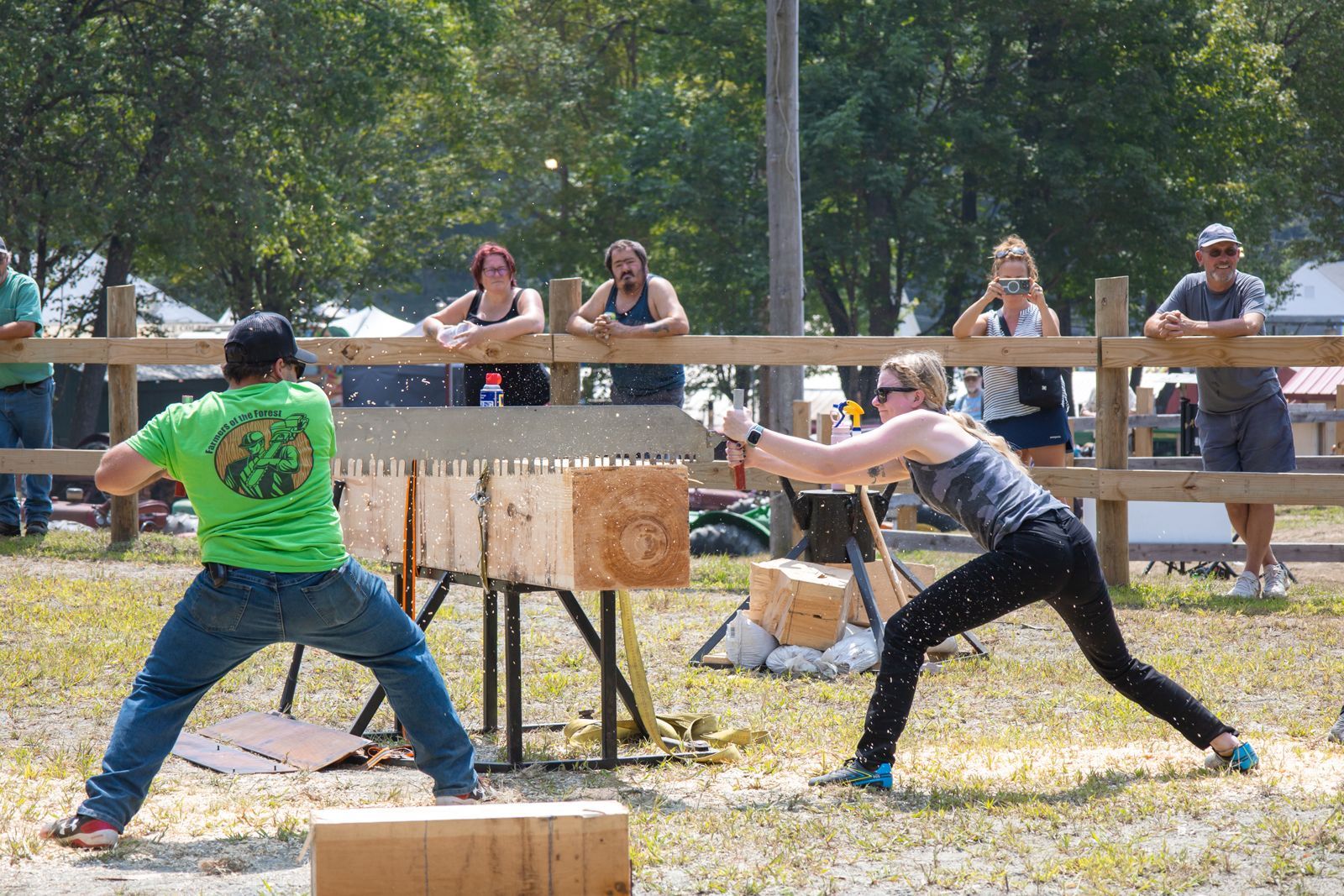 Two people swing axes at a log target outdoors, with spectators watching behind a wooden barrier.
