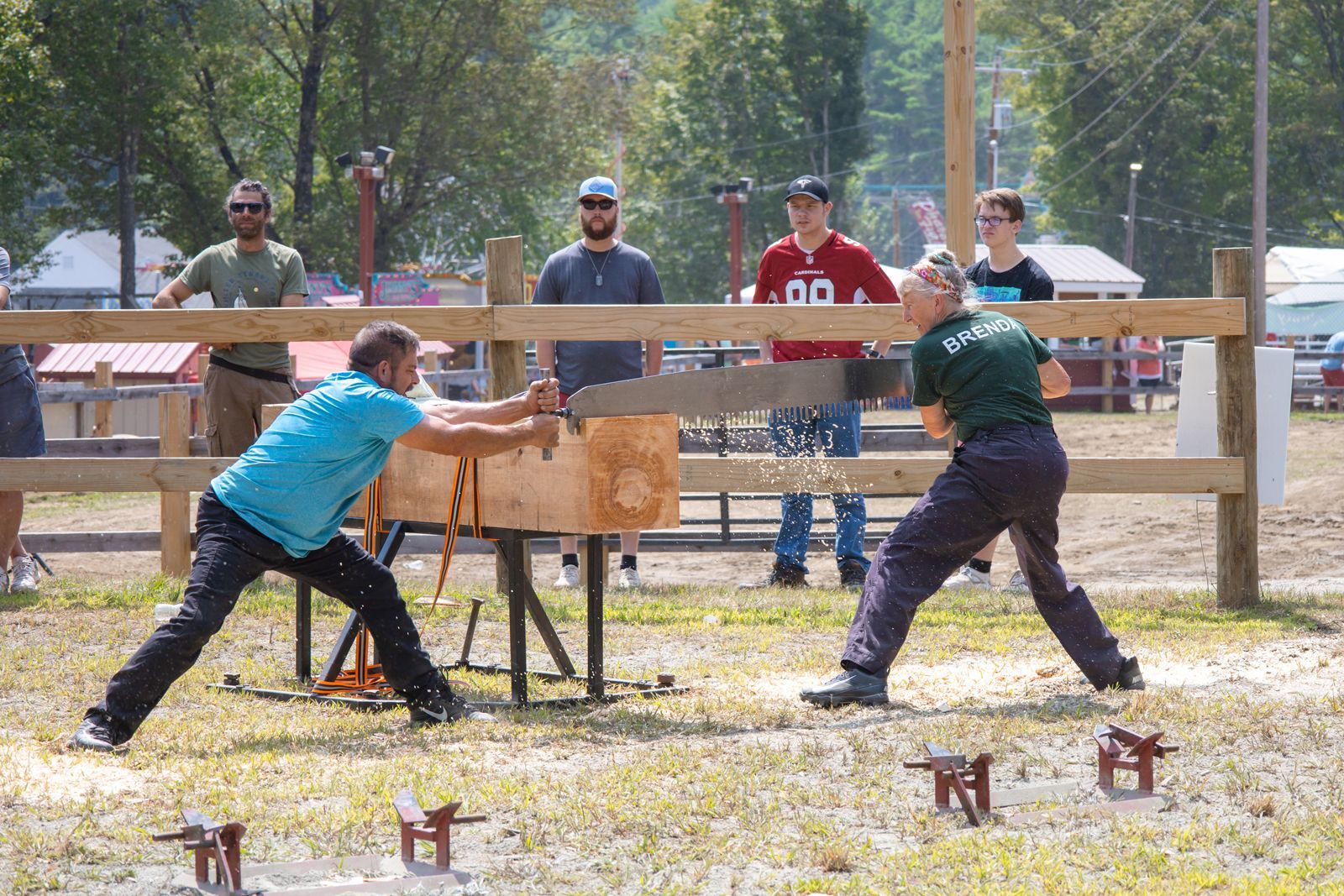 Two people swing axes at wooden targets in an outdoor lumberjack competition, with spectators behind a fence.