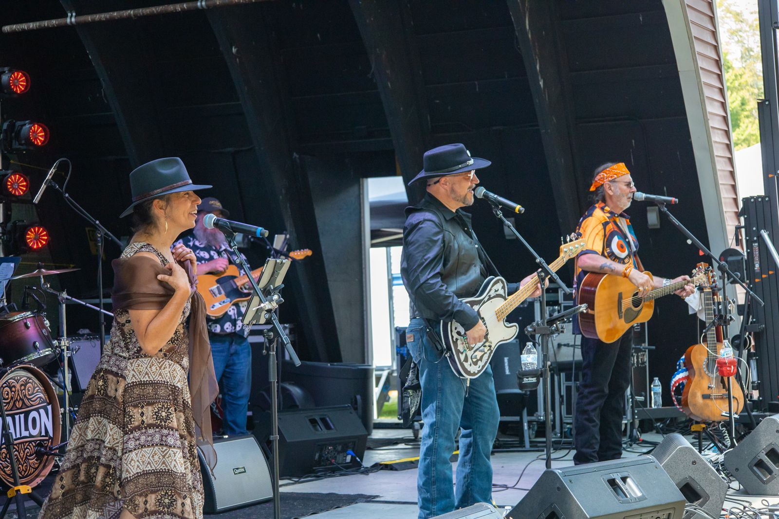 Country band performing on an outdoor stage, with guitars, microphones, and speakers under a canopy.